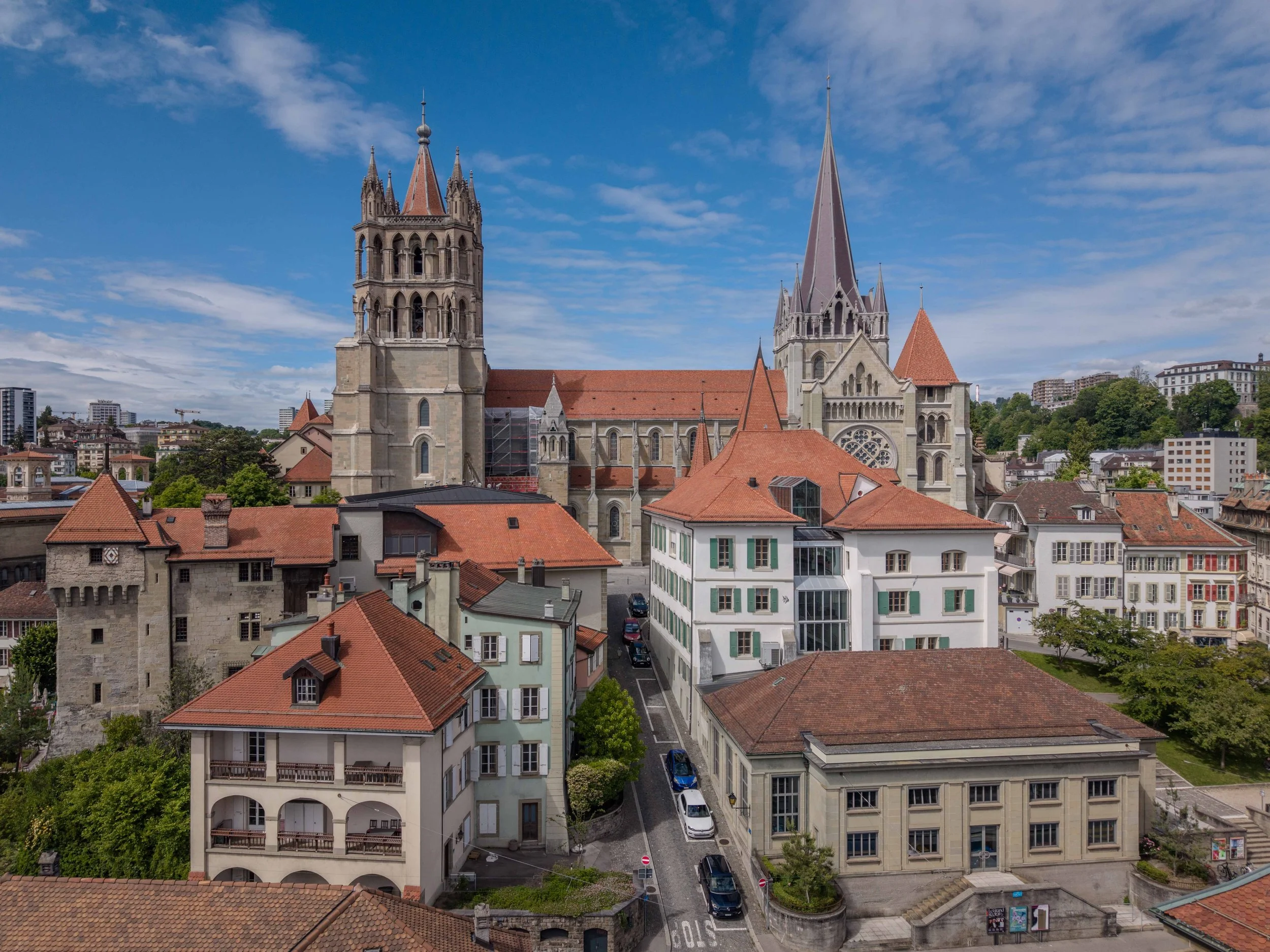 Vue aérienne de bâtiments anciens avec toits en tuiles rouges et deux tours d’église gothiques, sous un ciel partiellement nuageux.