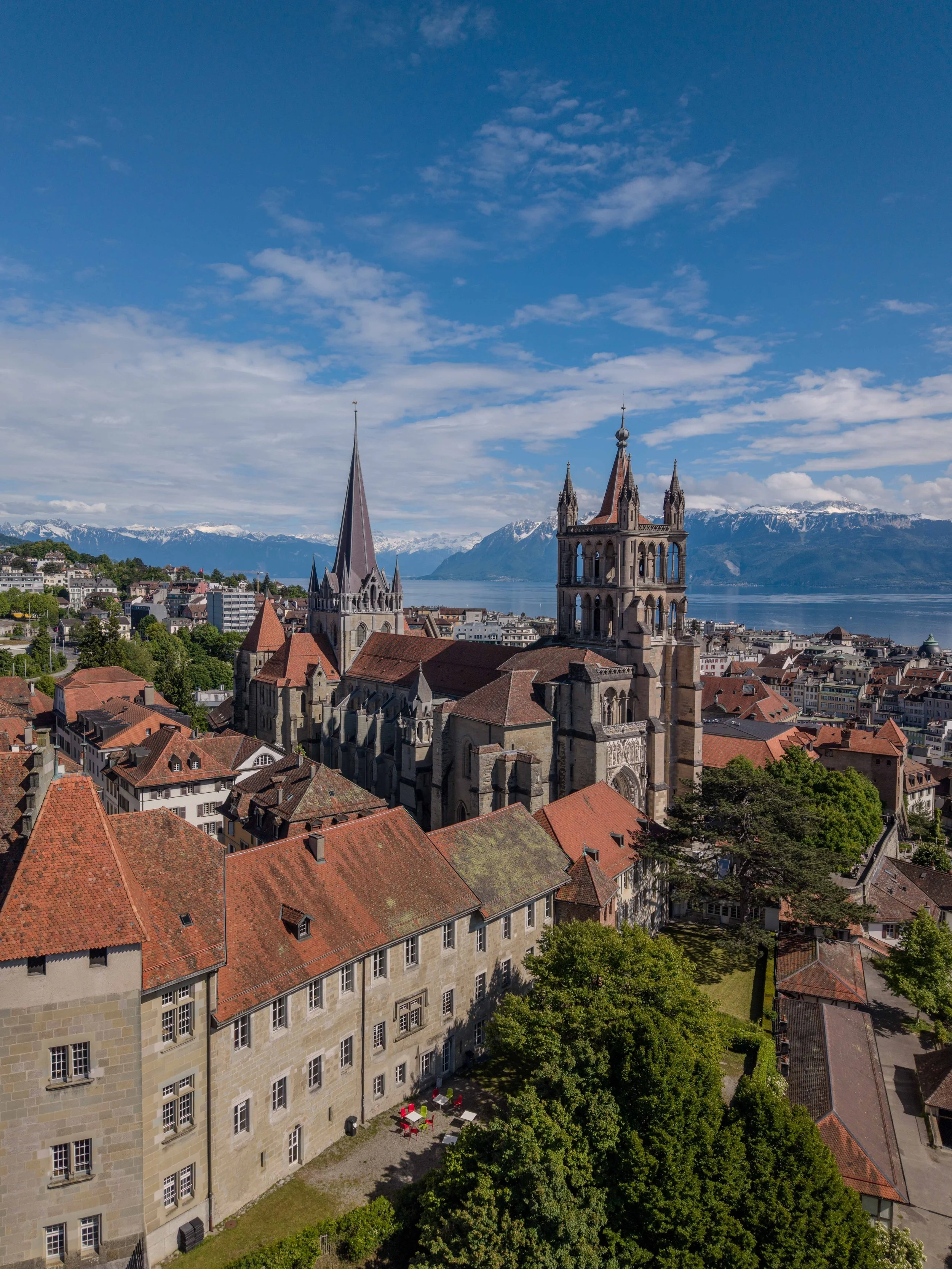 Vue aérienne d'une vieille ville avec des bâtiments en pierre, des toits en tuiles rouges, deux clochers d'église et des montagnes enneigées en arrière-plan, avec un ciel bleu et quelques nuages.