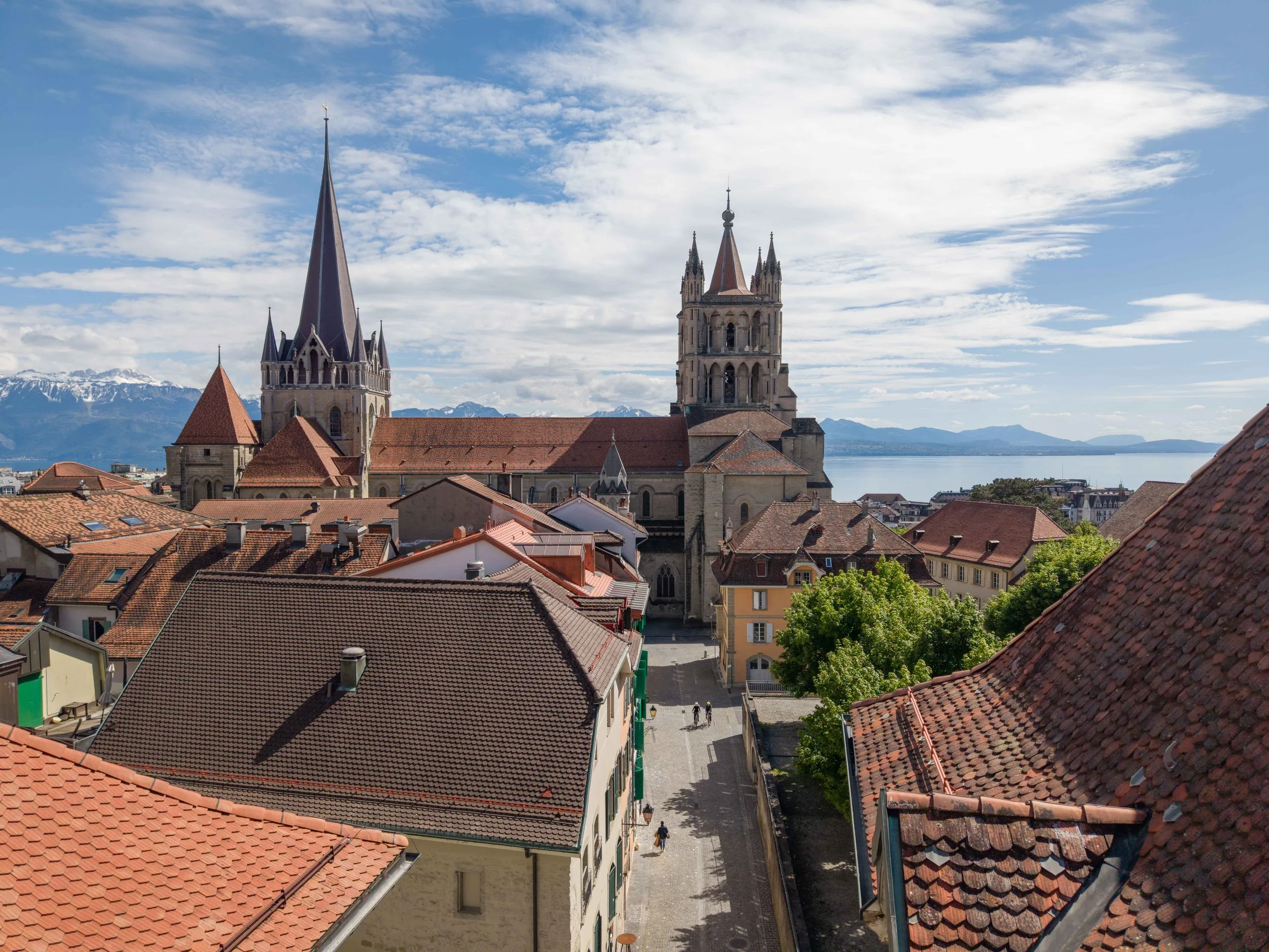 Une ville avec des toits en tuiles rouges, des églises avec des flèches pointues, et un lac avec des montagnes en arrière-plan sous un ciel partiellement nuageux.
