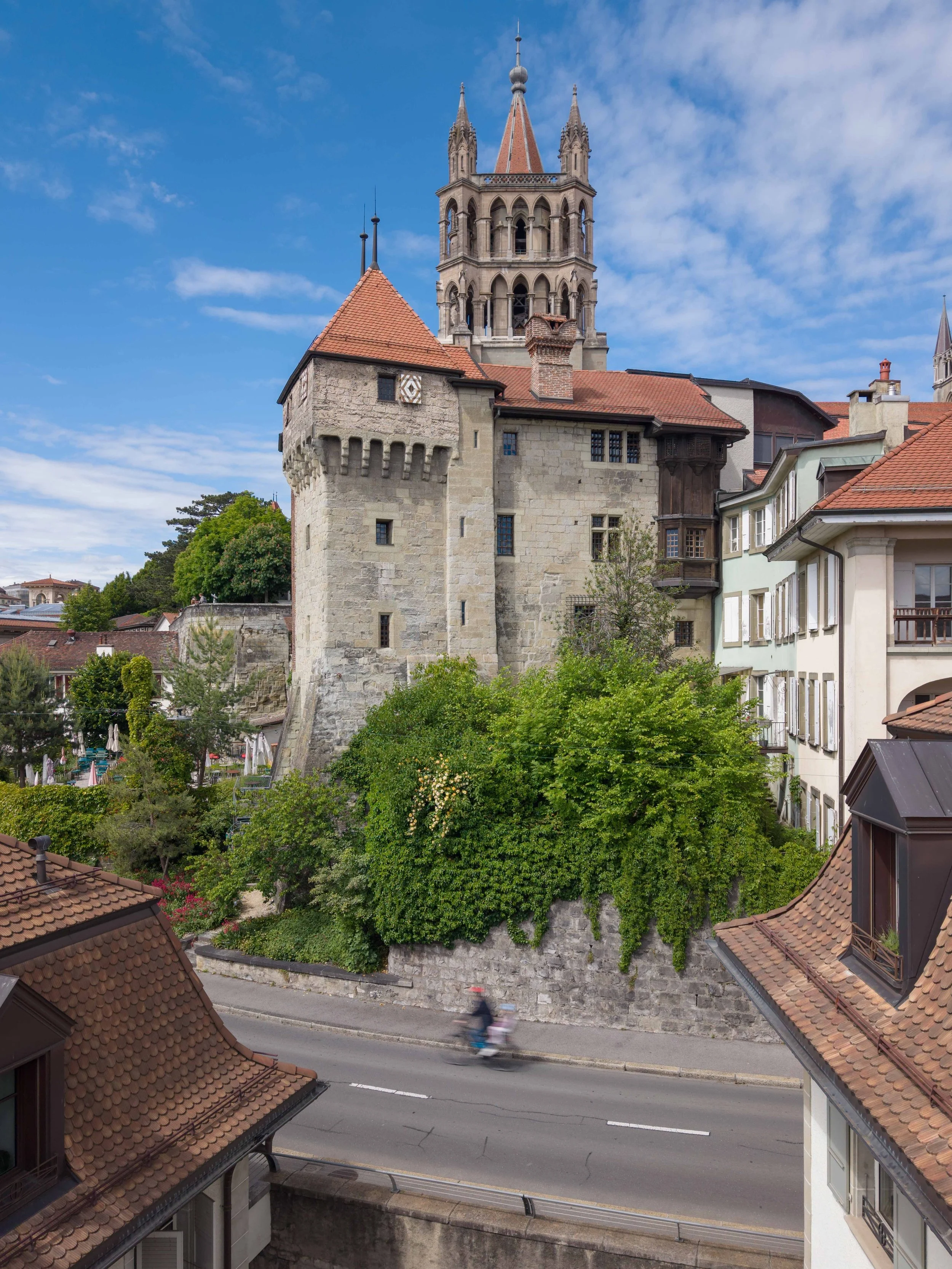 Vue d'une vieille tour médiévale en pierre avec des fenêtres à arc et un toit en tuiles rouges, vue sur un ciel bleu avec quelques nuages, entourée de maisons et de végétation urbaine.