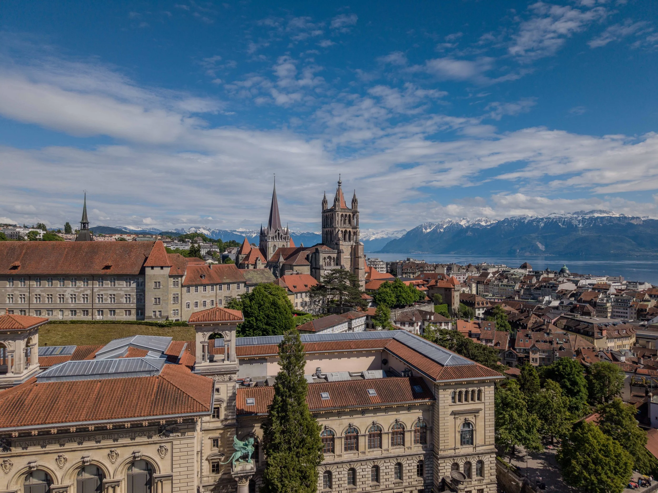 Vue aérienne d'une ville avec des bâtiments historiques en toiture rouge, des clochers d'églises, et un lac en arrière-plan avec des montagnes enneigées sous un ciel partiellement nuageux.