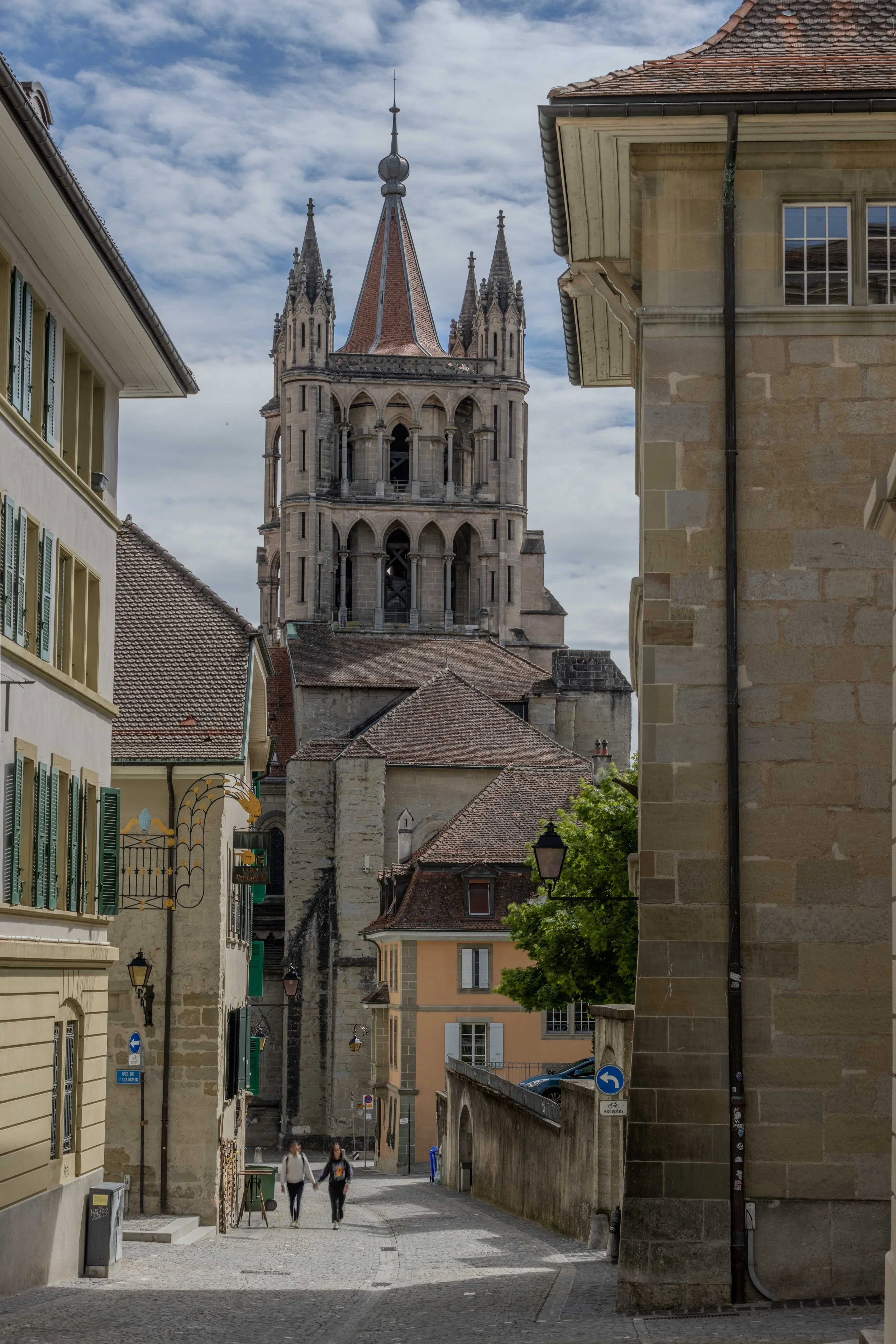 Vue d'une rue de ville avec des bâtiments anciens et une grande tour d'église en pierre au centre, ciel partiellement nuageux.