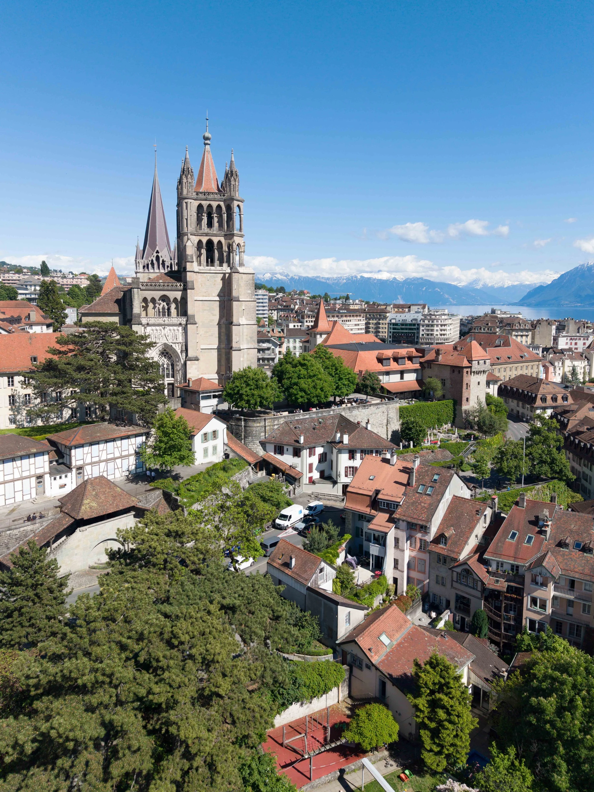Vue aérienne d'une ville ensoleillée avec des bâtiments historiques, un lac au loin et des montagnes avec sommets enneigés en arrière-plan.