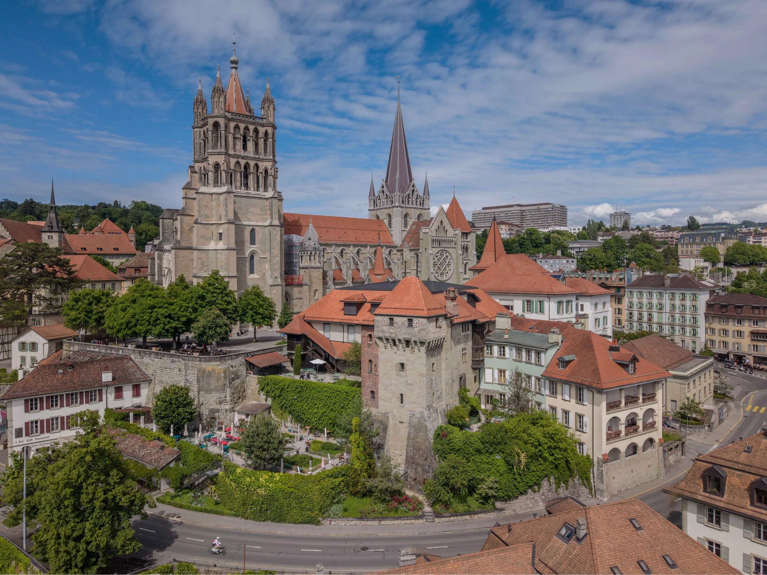 Vue aérienne d'une ville avec une architecture de style européen, comprenant des églises avec de grandes tours pointues, des bâtiments résidentiels, une rue avec des cyclistes, des arbres verts et un ciel partiellement nuageux.