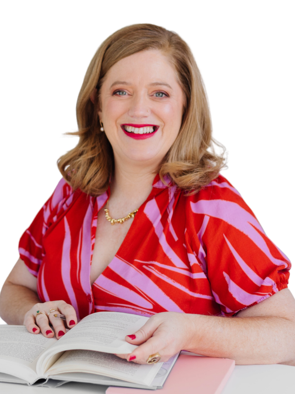 A woman with shoulder-length red hair, smiling, wearing red lipstick, a pearl necklace, and a red, pink, and white patterned dress, sitting at a table with an open book.