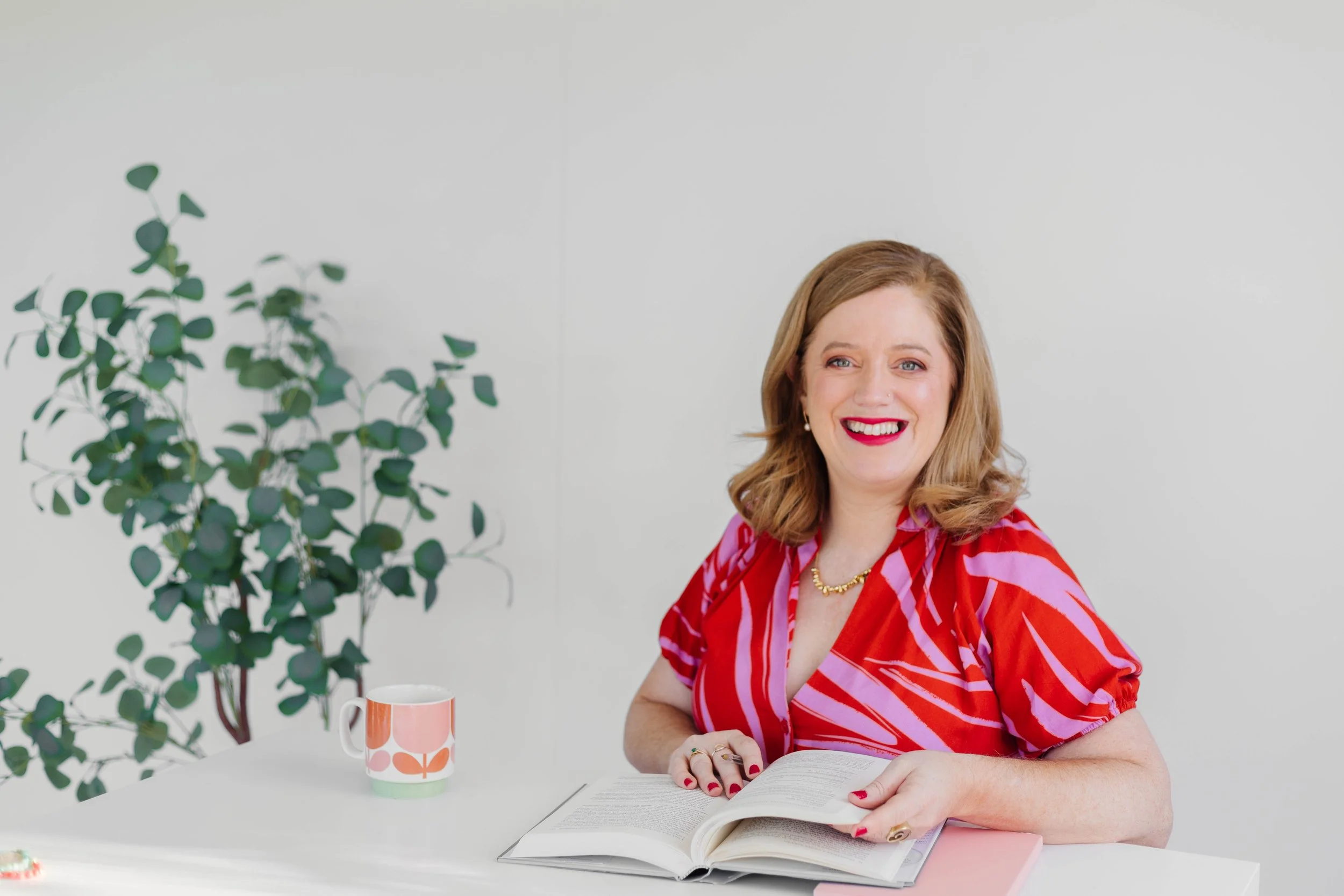 A woman with shoulder-length light brown hair, wearing a red and pink striped blouse, smiling while sitting at a white desk with an open book, a pink notebook, a colorful mug, and a green leafy plant against a plain light background.
