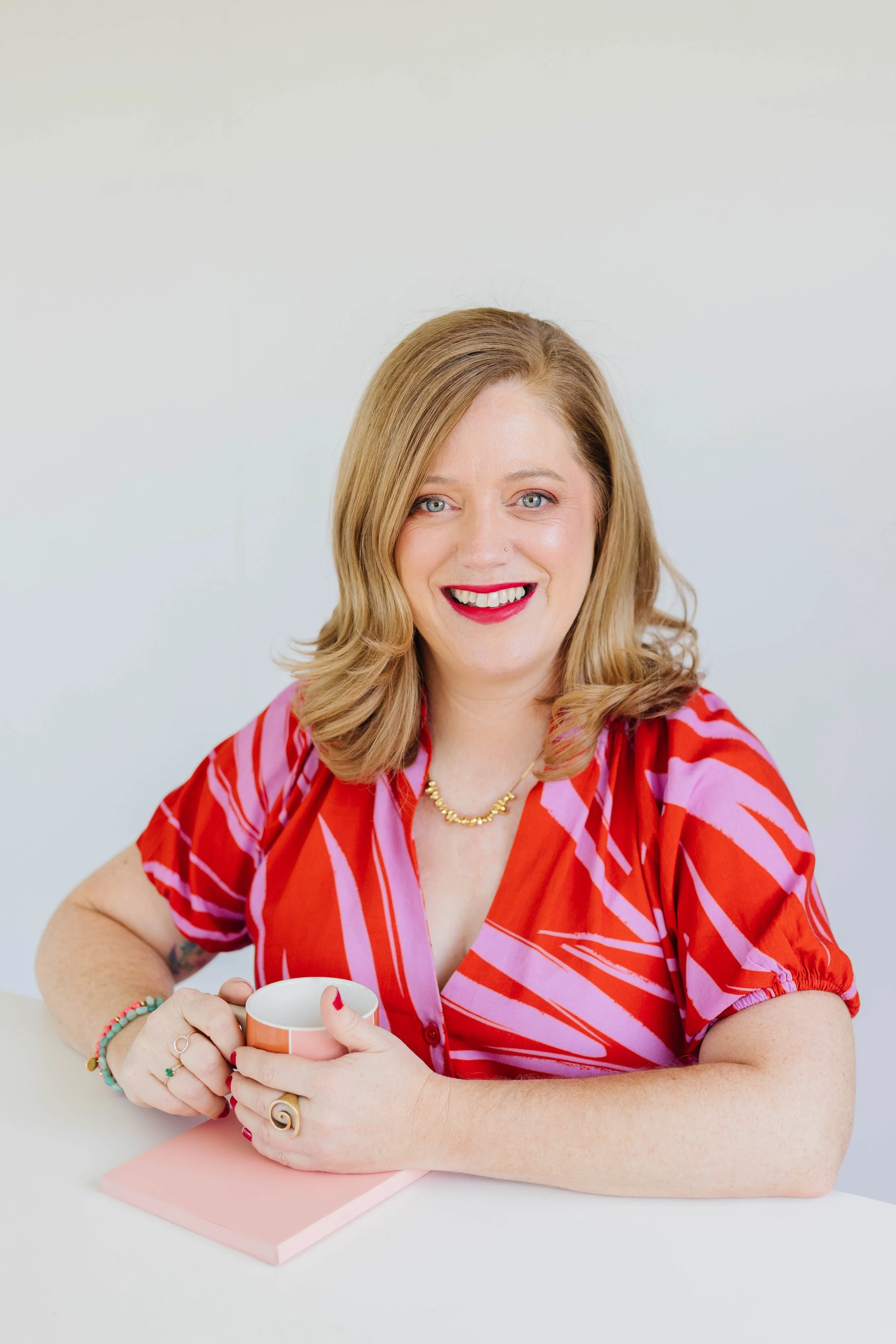 A woman with blonde hair, wearing a red and pink patterned shirt, smiling, holding a mug, and sitting at a white table with a light pink notebook.