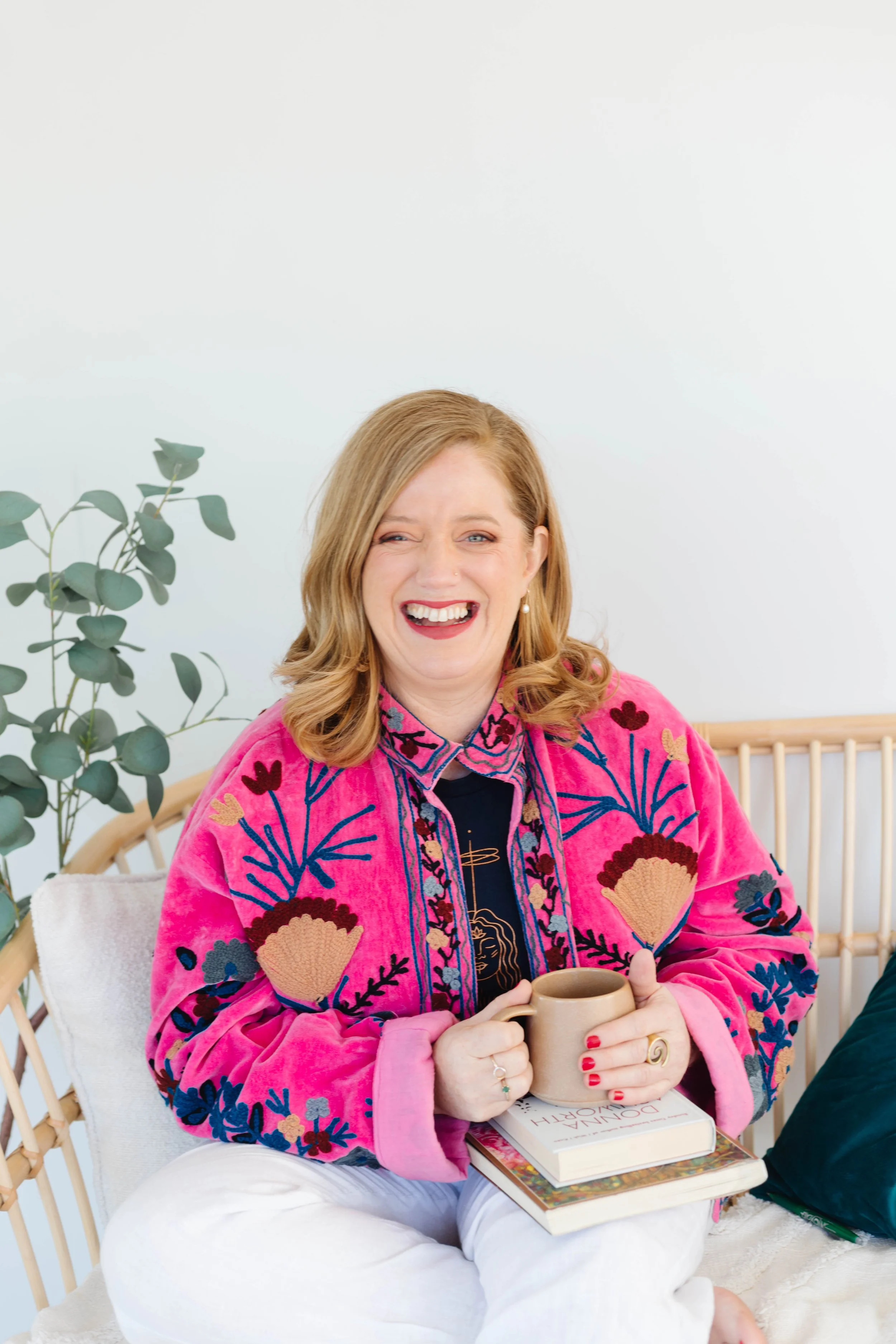Woman with red hair smiling, wearing a bright pink embroidered jacket, holding a beige mug, sitting on a white chair with books and a plant nearby.
