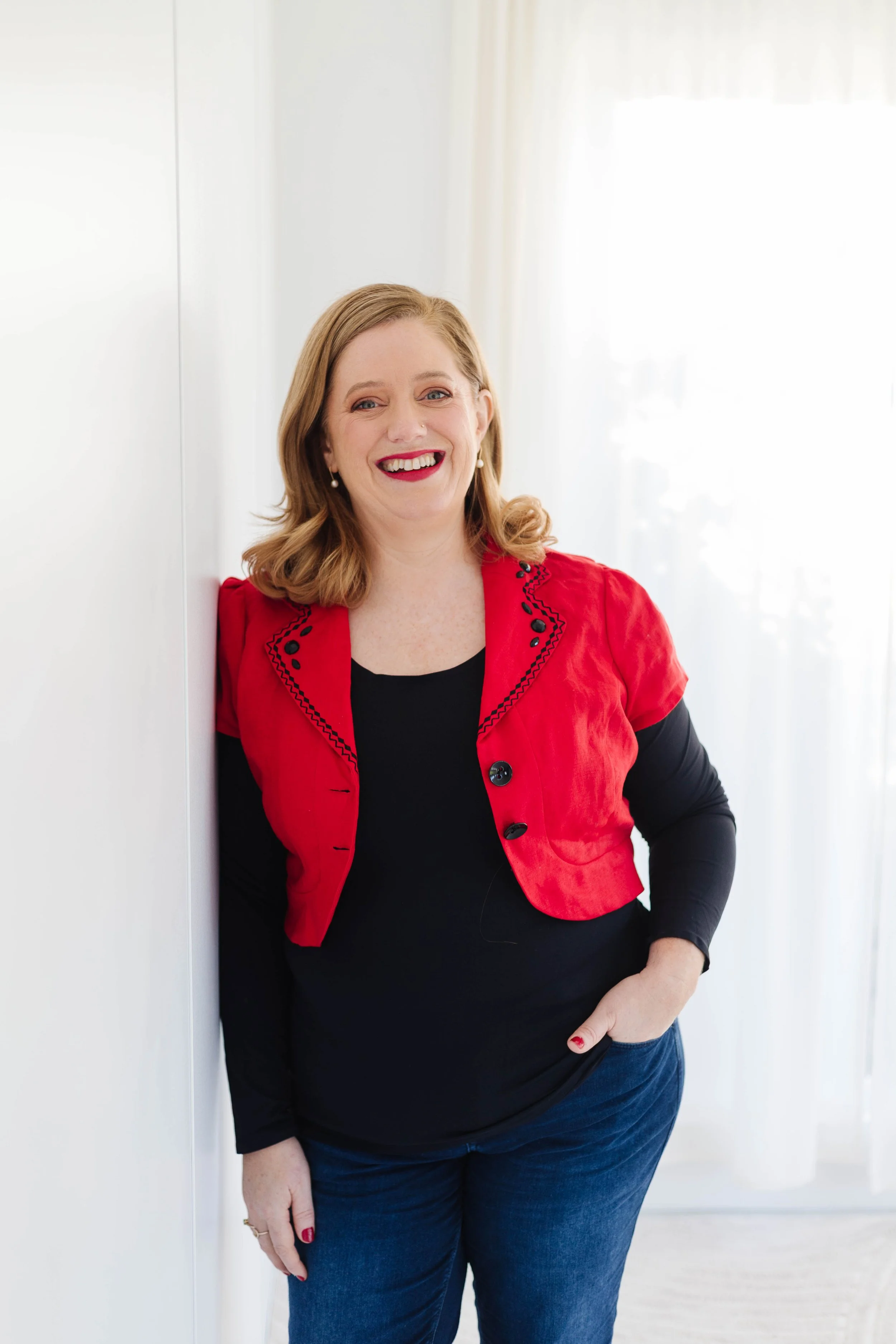A woman with red hair wearing a red jacket over a black top and blue jeans, smiling and leaning against a white wall near a window with sheer curtains.
