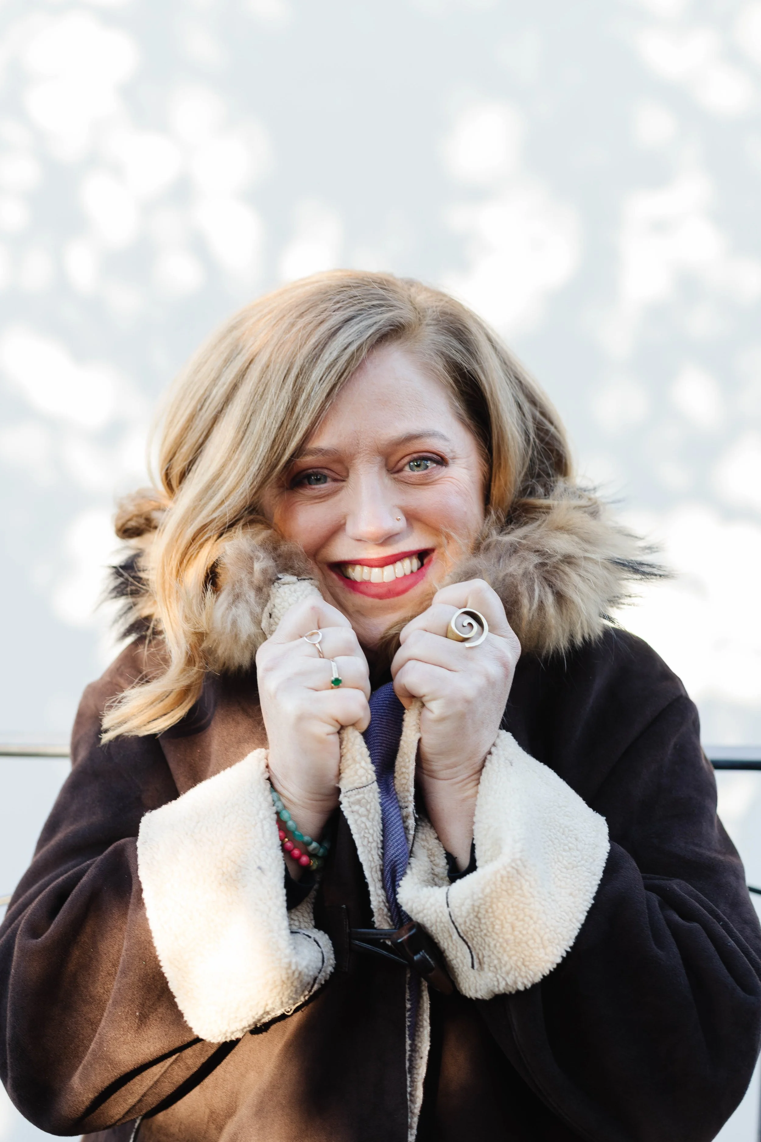 A woman with blonde hair, smiling and wearing a coat with a fur-lined hood, stands outside with a blurred background of leafless trees and sky.