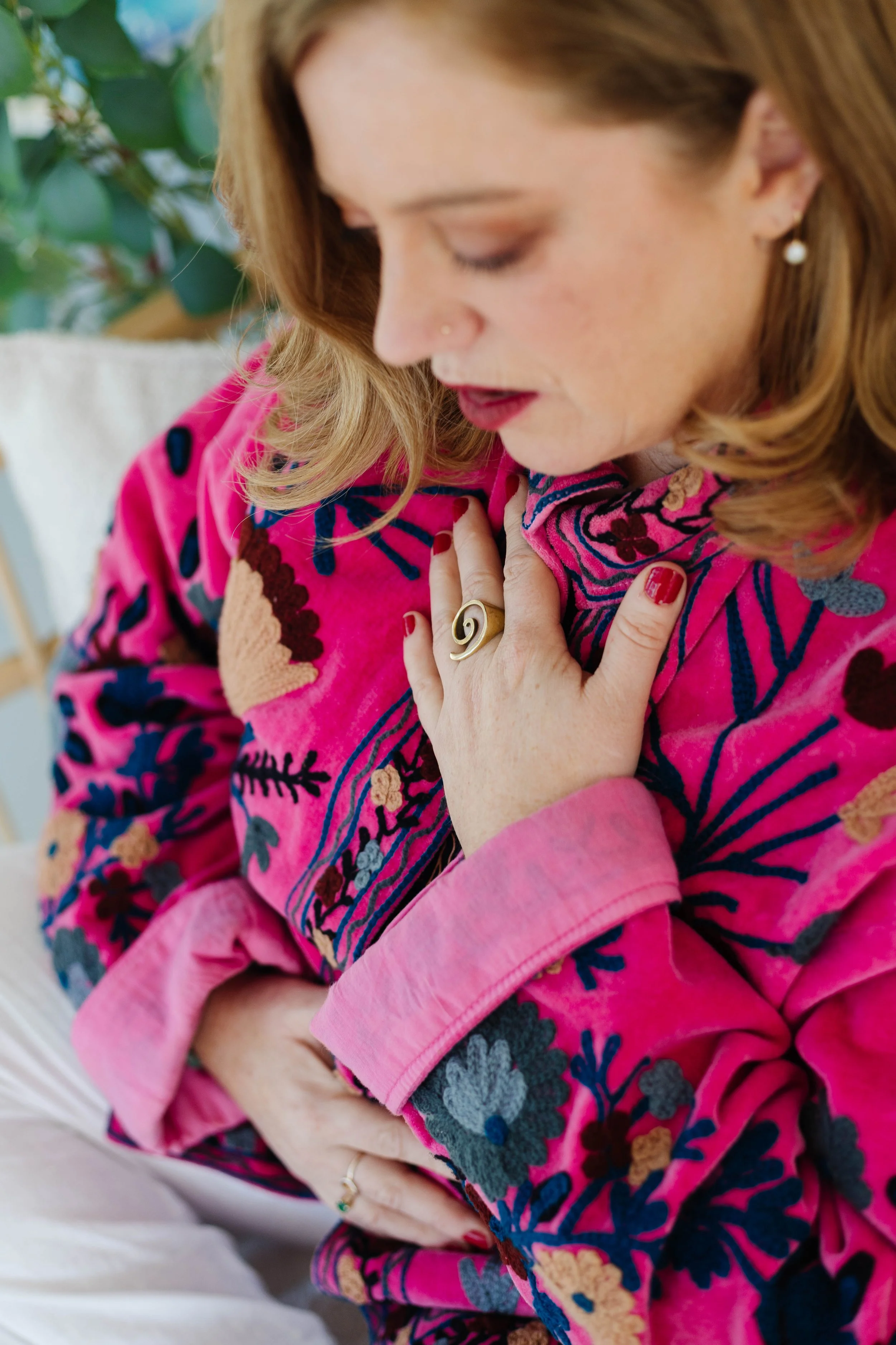 A woman with red hair and a nose ring wearing a vibrant pink floral embroidered jacket, touching her chest with one hand, and resting her other hand on her stomach, wearing rings and nail polish.