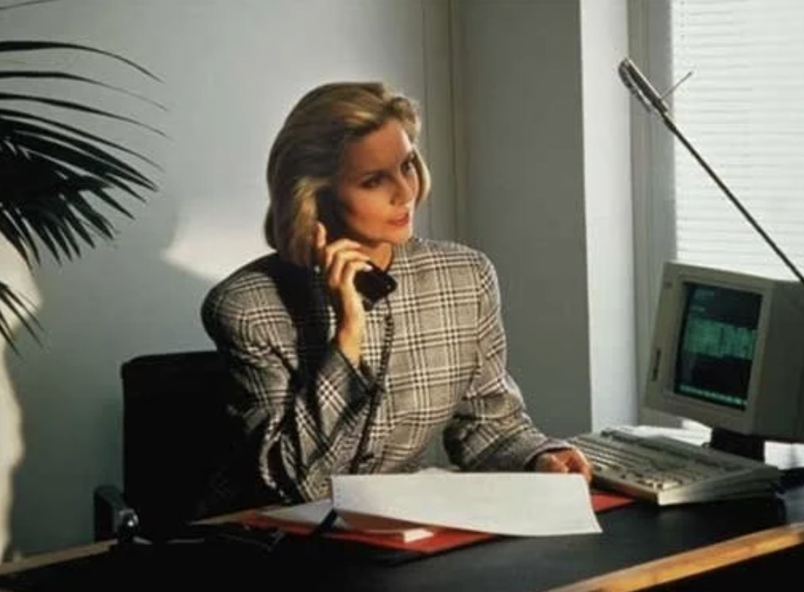 A woman wearing a checkered blazer sitting at an office desk, talking on a landline phone, with papers, a computer monitor, keyboard, and a lamp on the desk.