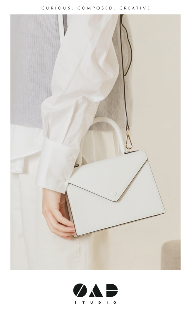 Close-up of a person holding a white handbag with a black strap, wearing a white long-sleeve shirt and a light gray sweater, with text "Curious, Composed, Creative" at the top and "OAD Studio" at the bottom.