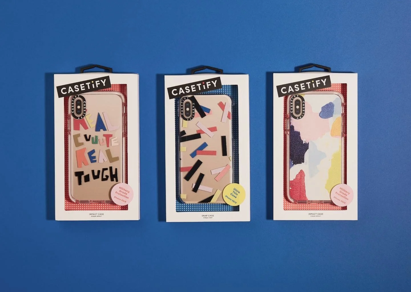 Three iPhone cases in packaging on a blue background. The first case has colorful letters spelling 'MAKE EVERYDAY KID TUGH', the second has abstract shapes in pink, black, blue, and yellow, and the third features an abstract watercolor pattern with pastel colors.