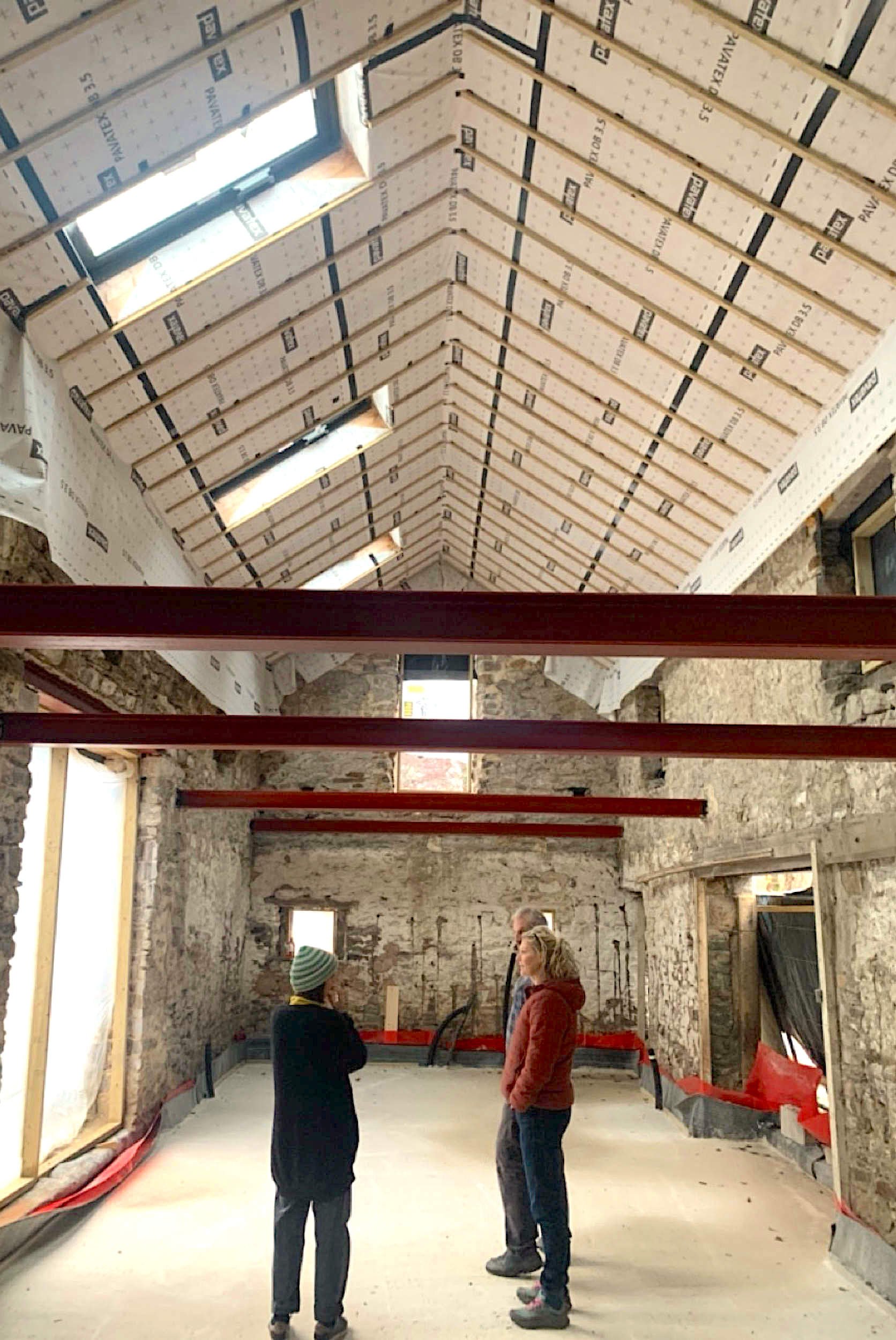 Interior of a building under construction, showing exposed stone walls, a pitched roof with insulation and skylights, and three people standing and talking on a concrete floor.