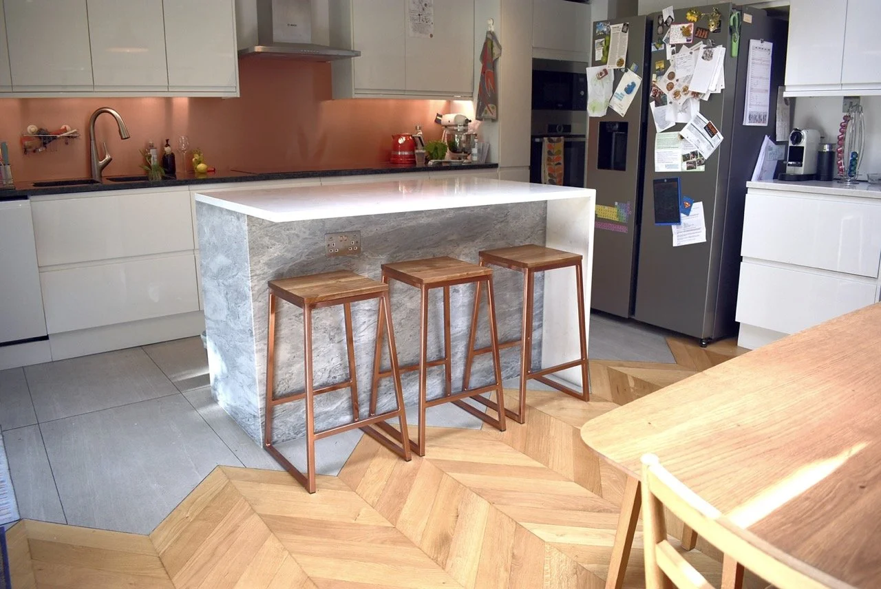 Modern kitchen with a marble island, three wooden bar stools, white cabinets, a stainless steel refrigerator covered in magnets and notes, and various appliances and decorations.