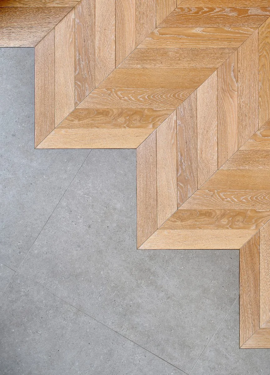 Close-up of a corner of a room featuring wooden parquet flooring in a herringbone pattern and large gray concrete tiles.