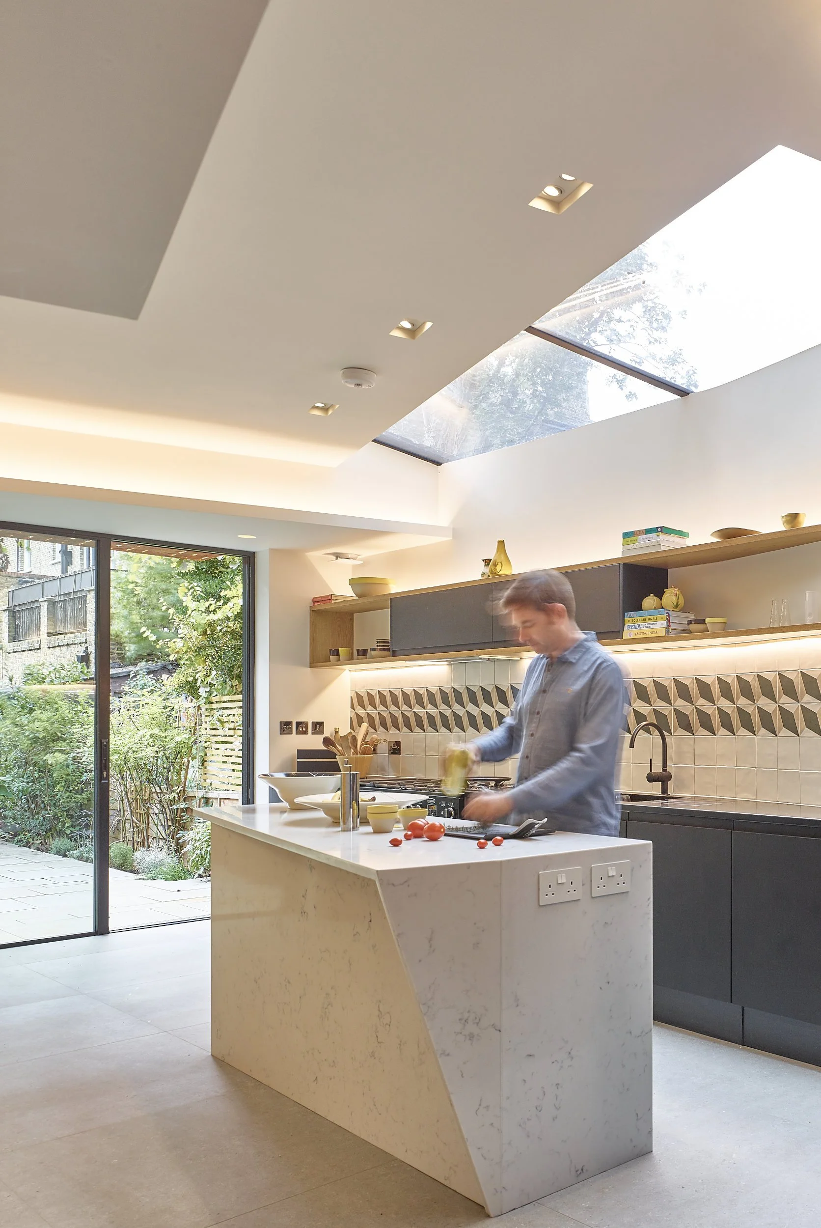 A person preparing food in a modern kitchen with a white marble island, open shelving with dishes and books, large sliding glass doors leading to a garden, and a skylight to let in natural light.