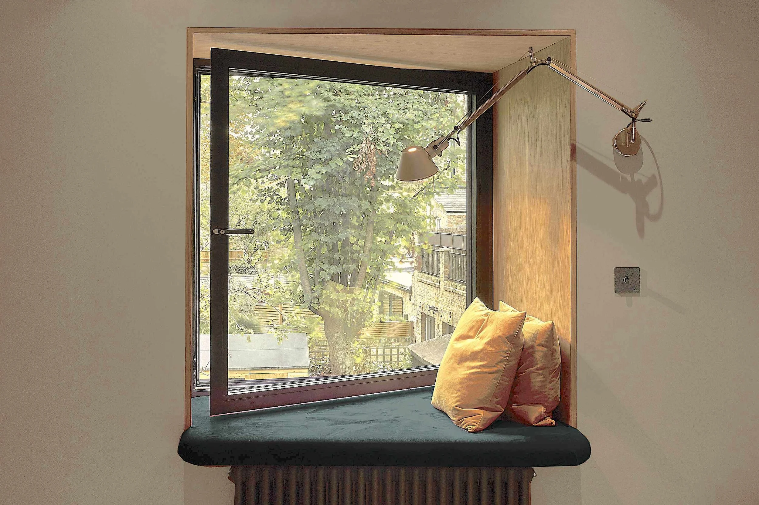 A window with a wooden frame showing trees outside, with sunlight filtering through the leaves. Inside, there is a corner with a wall-mounted adjustable lamp casting a shadow, a beige cushion on a black surface, and a dark wooden base or furniture below.