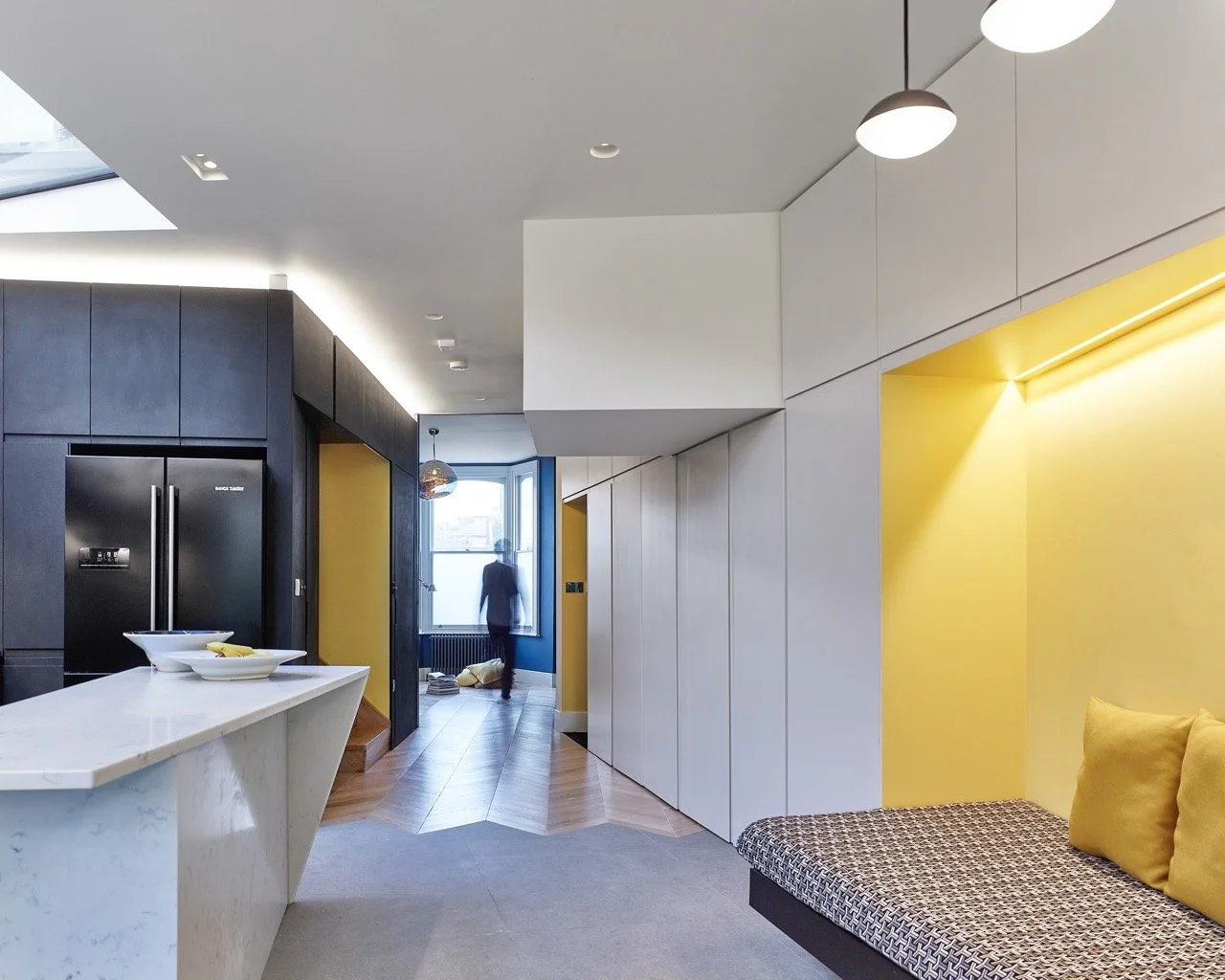 Modern apartment interior with black and white walls, yellow accent wall, marble kitchen island, and seating area with patterned bench and yellow pillows.