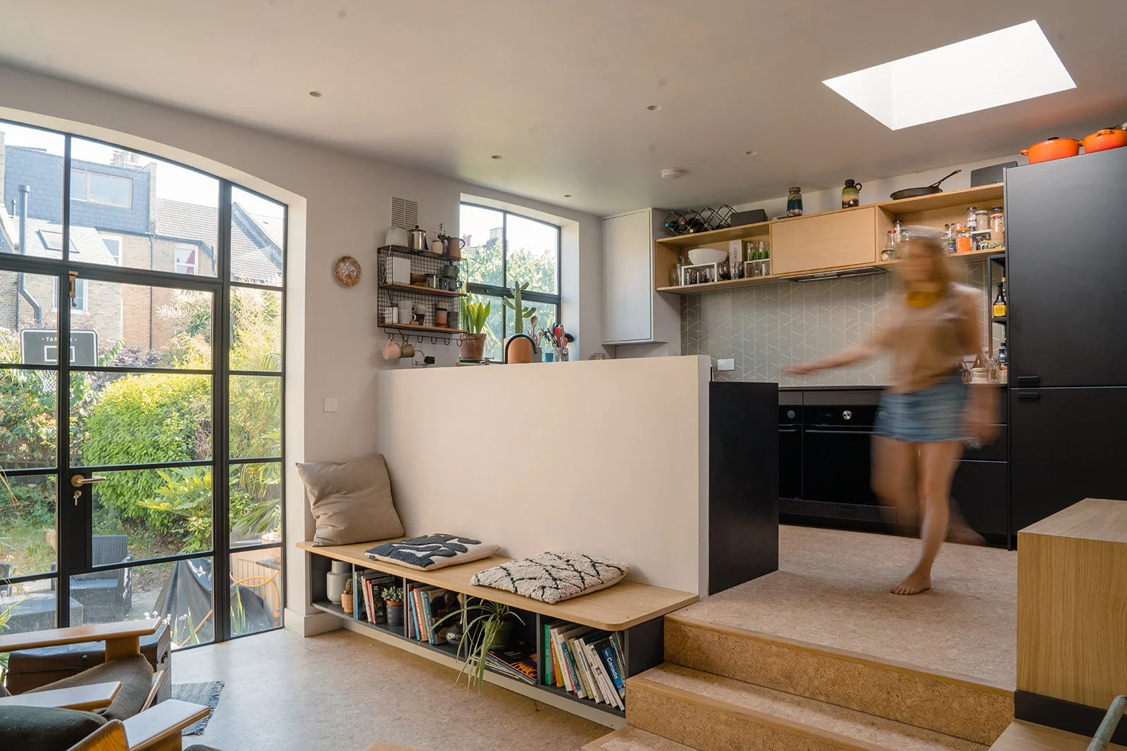 Bright, modern living room with large glass door, view of garden, to the left, a built-in shelf with books and cushions, and to the right a kitchen with wooden shelves, black cabinets, and a blurred figure of a woman walking.