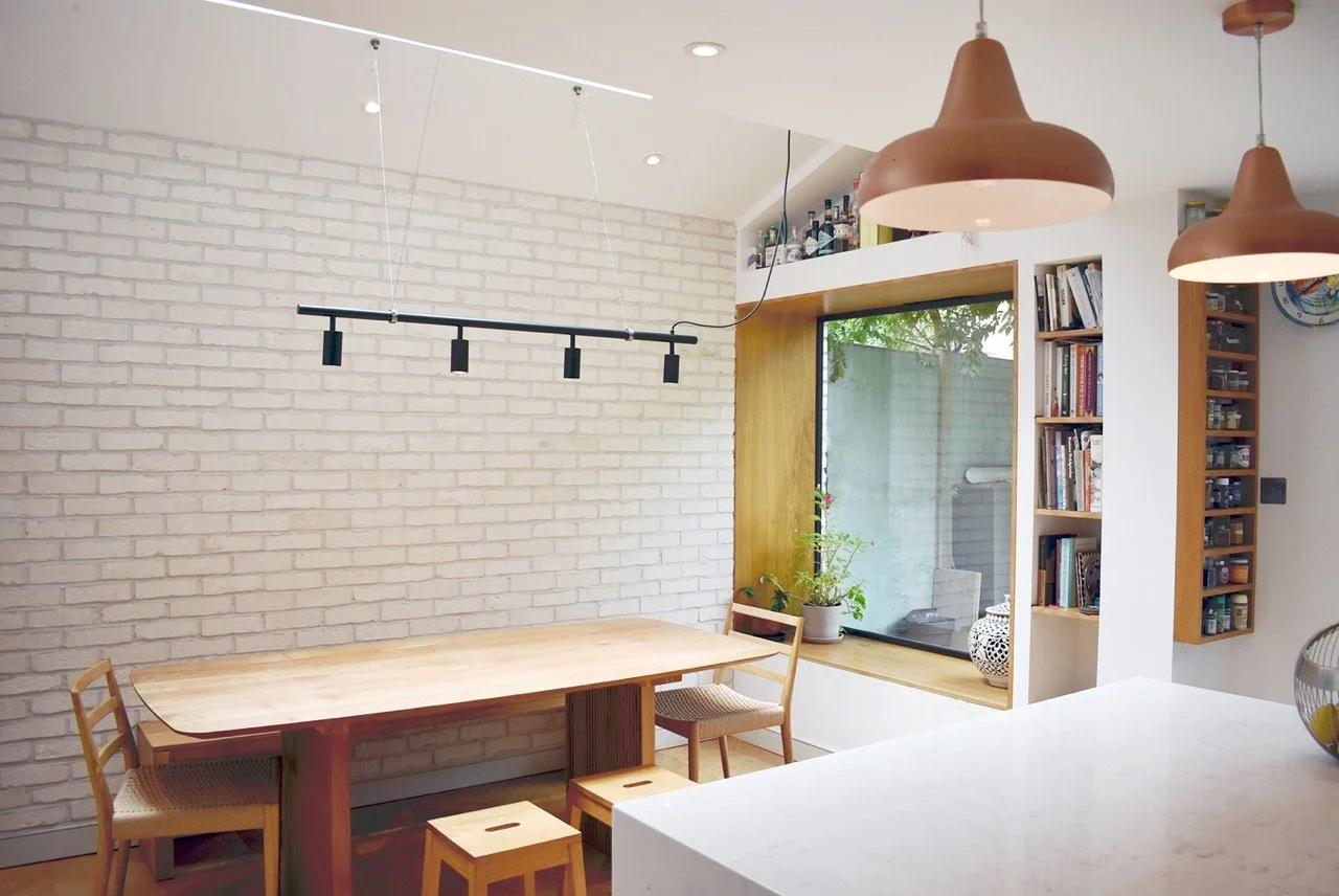 Modern dining area with a wooden table and chairs, a window seat with plants, and built-in bookshelves, in a room with white brick walls and hanging pendant lights.