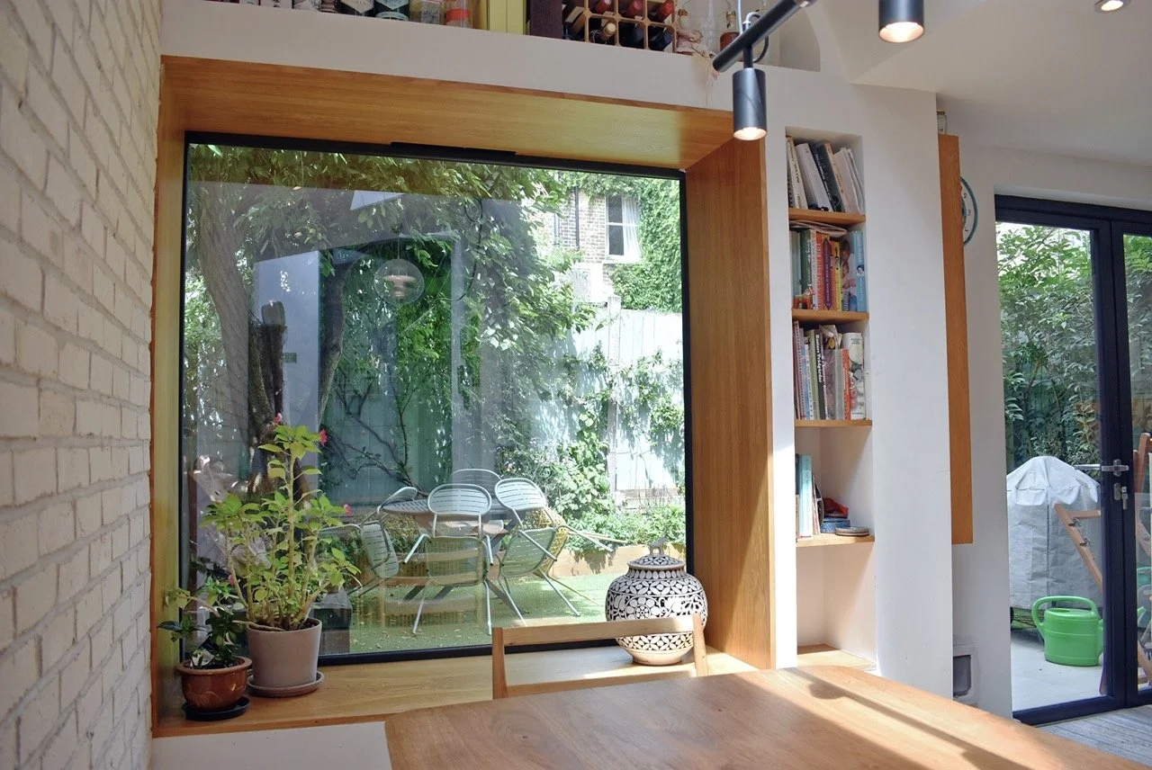 Interior of a modern home with a large window seat with oak surround, looking out onto a garden with chairs and trees, bookshelf on the right, potted plants on the left, and a wooden table in the foreground.