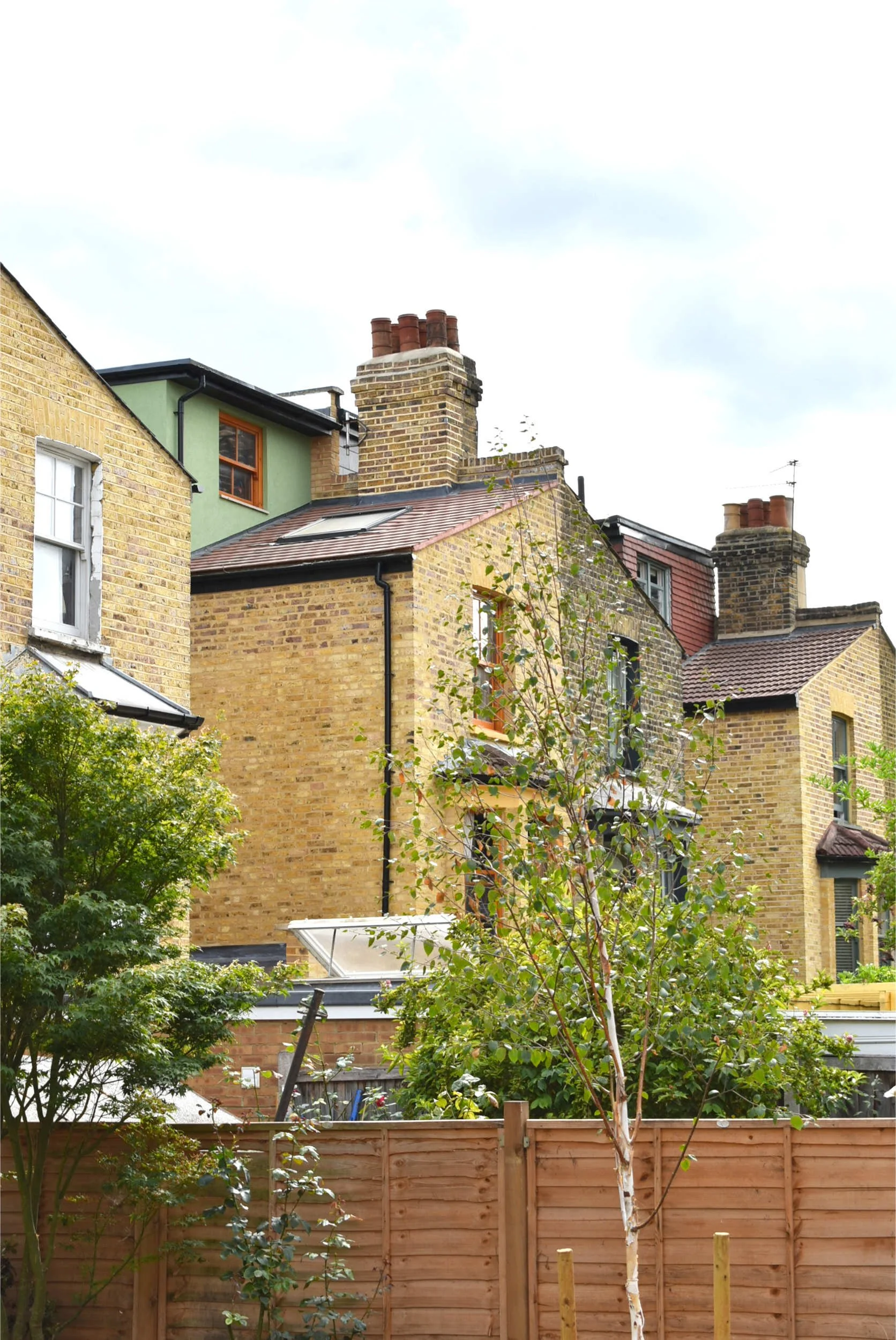 View of the rear of victorian terraced houses with brick facades, some with greenery and a wooden fence in the foreground.