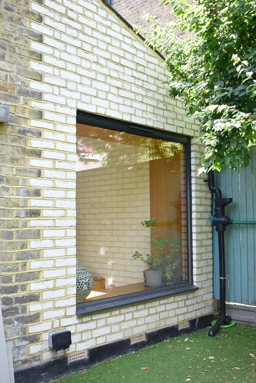 Photo of a house exterior with a contemporary window seat, brick walls, a green fence, and a tree with green leaves. Inside the window, a potted plant and decorative objects are visible.