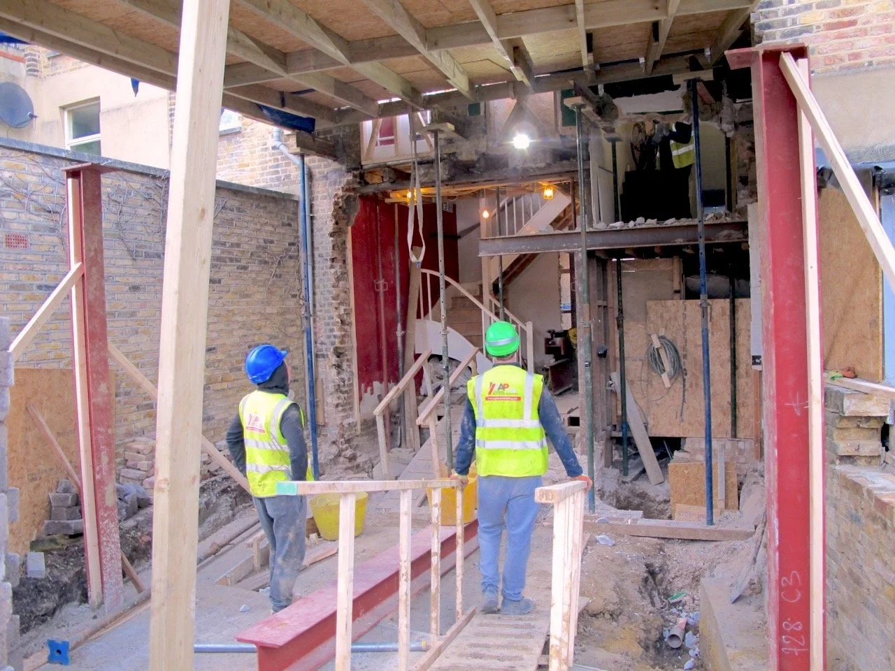 Construction workers in safety vests and helmets at building site with exposed brick walls and scaffolding.
