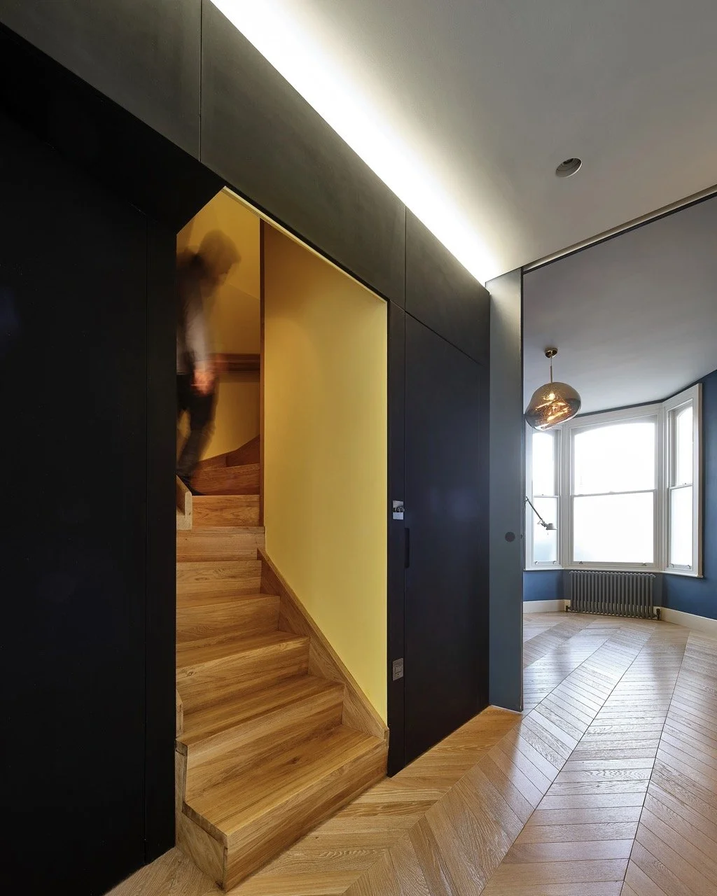 Interior of a modern house featuring a flight of wooden stairs with a yellow wall, a neighboring room with blue walls, large windows, and wooden flooring.