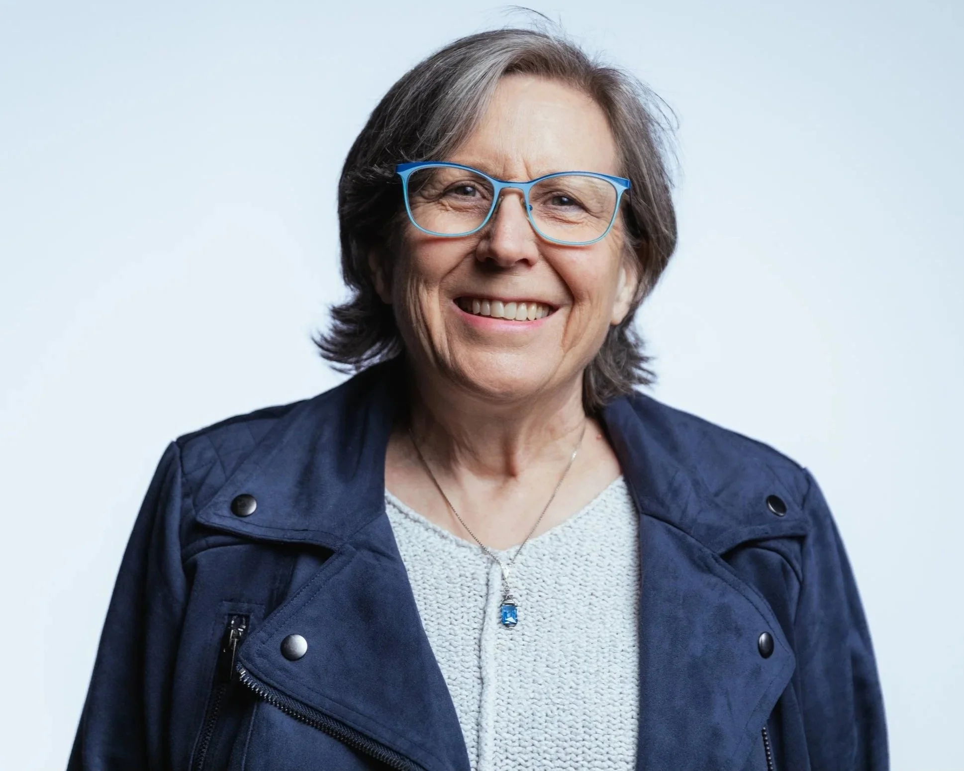 An older woman wearing blue glasses, a navy jacket, and a light-colored sweater, smiling against a plain light background.