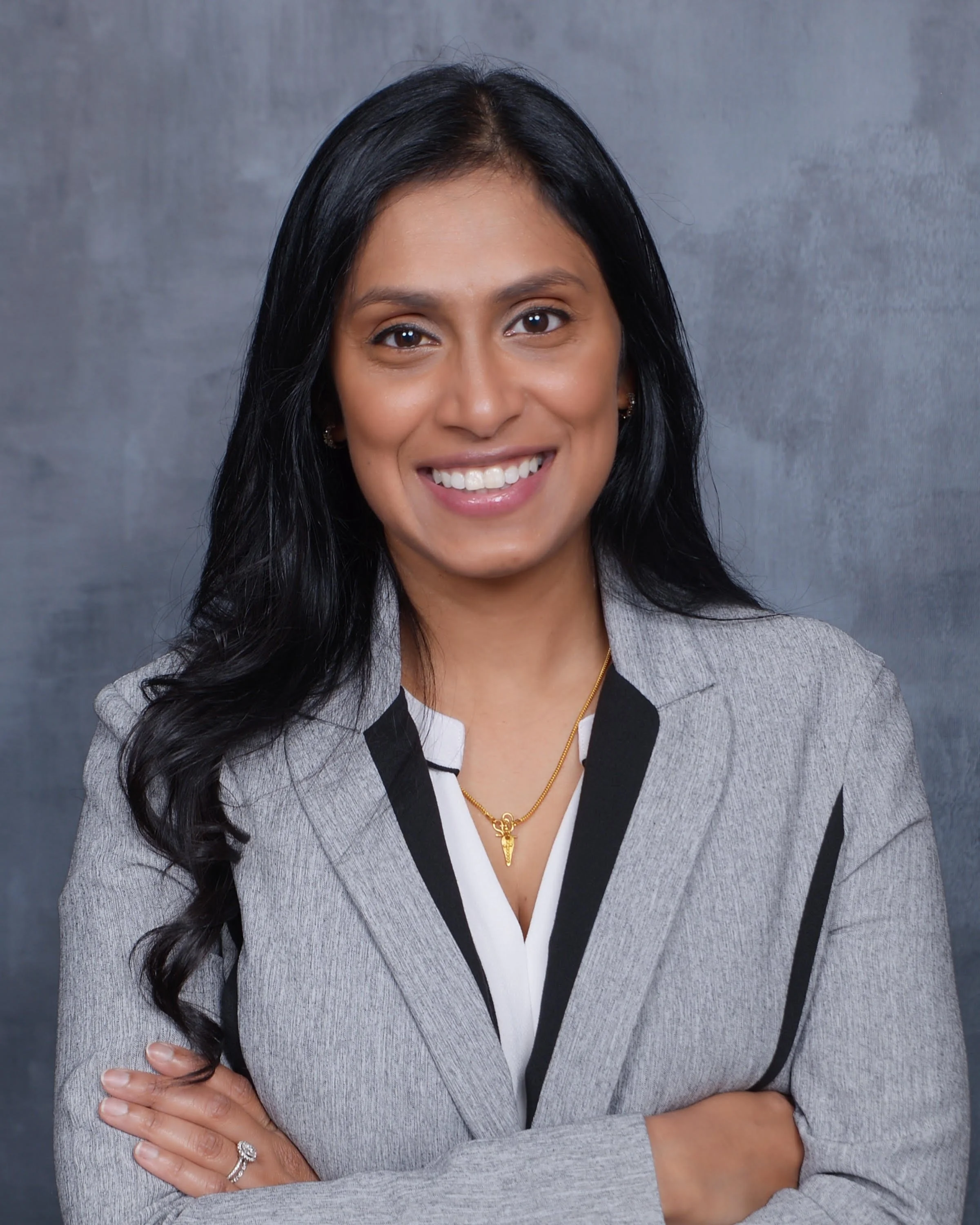 A woman with long black hair smiling, wearing a gray blazer with black accents, a white shirt, a gold necklace, and rings, standing against a gray background.