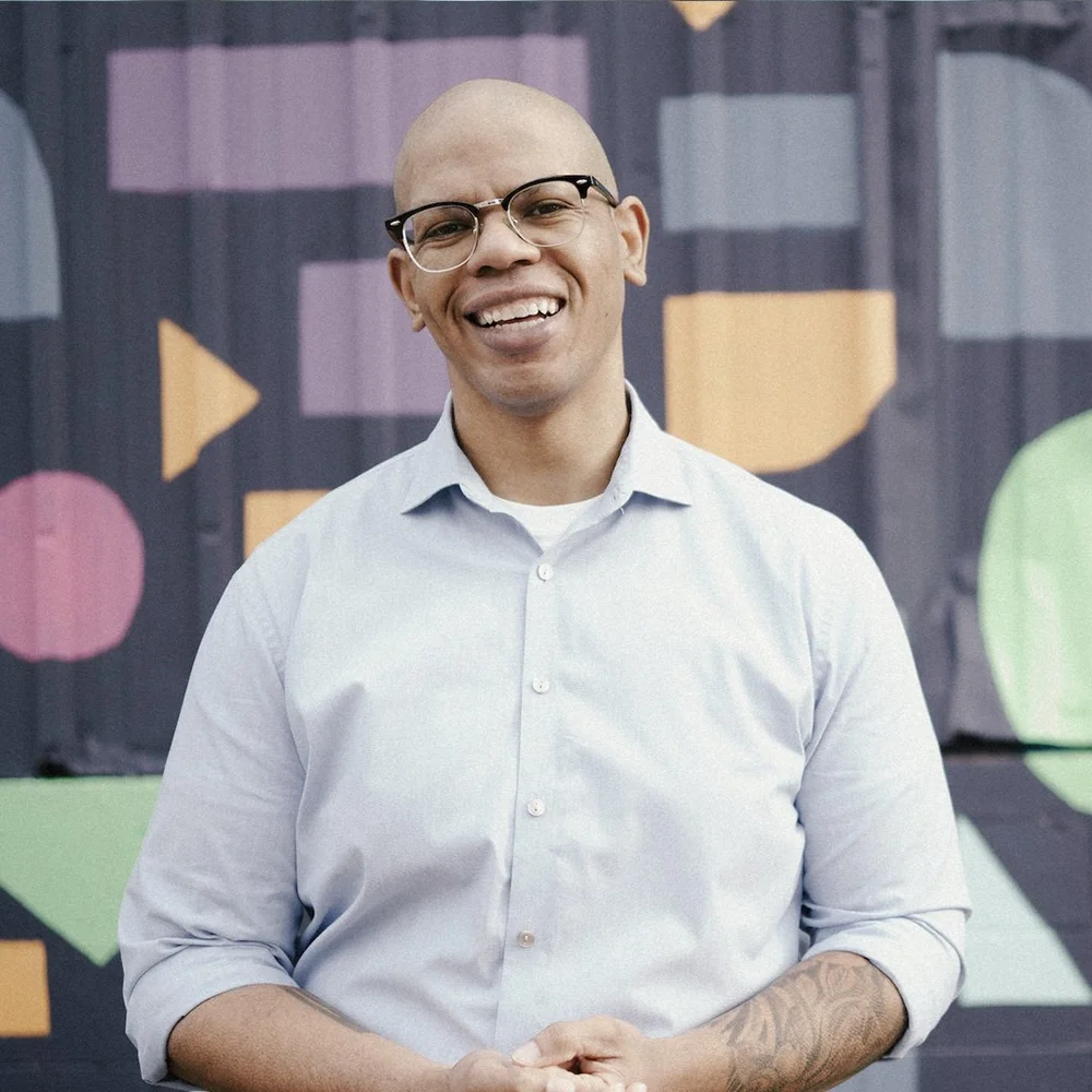 A smiling bald man with glasses wearing a light blue button-up shirt standing in front of a colorful geometric-patterned backdrop.