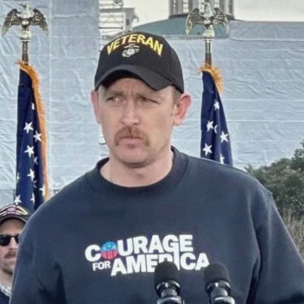 Pete Lucier wearing a veteran cap in front of American flags.