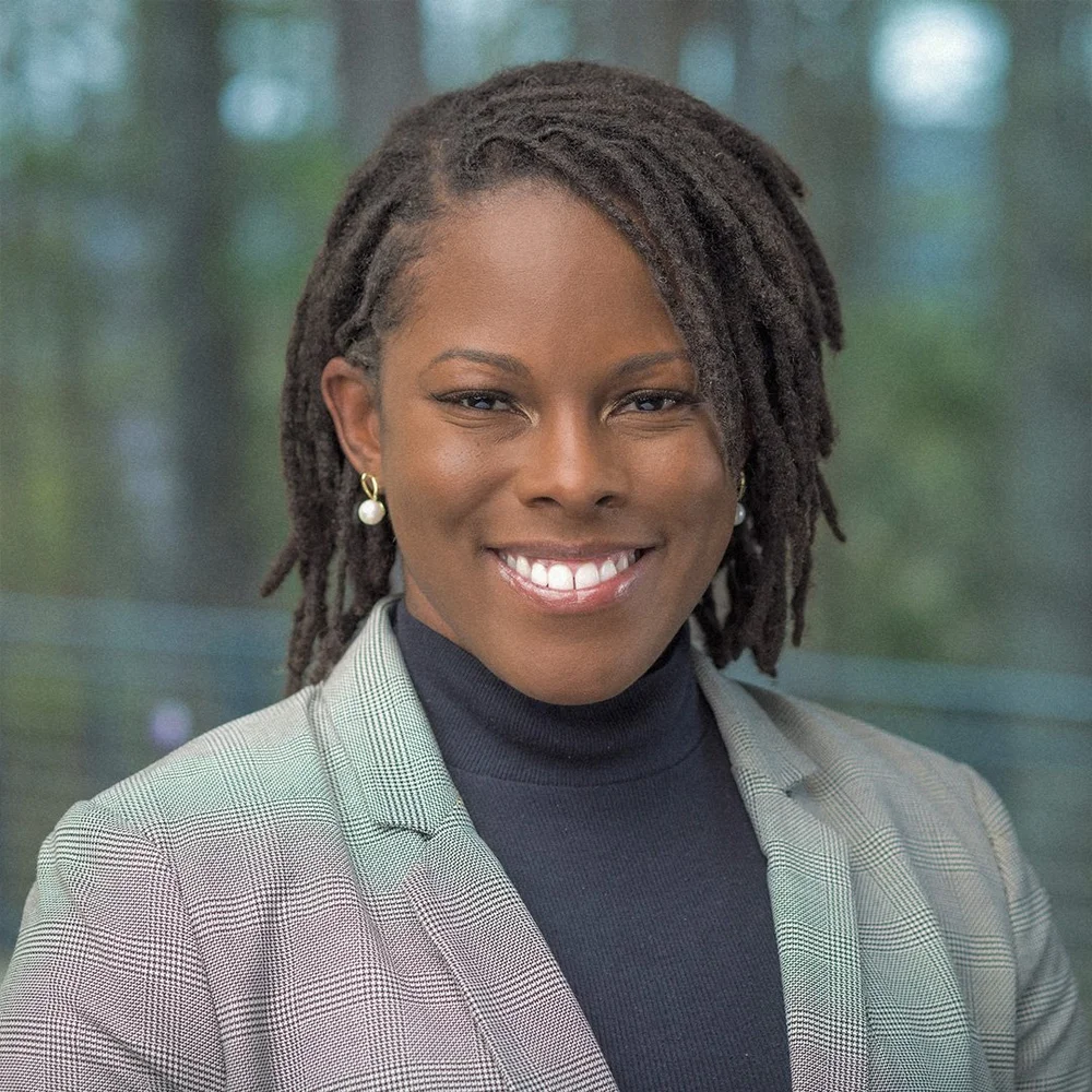 Close-up of a smiling woman with dark skin, wearing a gray plaid blazer, black turtleneck, gold earrings with pearls, and dreadlocks, in front of a green, blurred background.