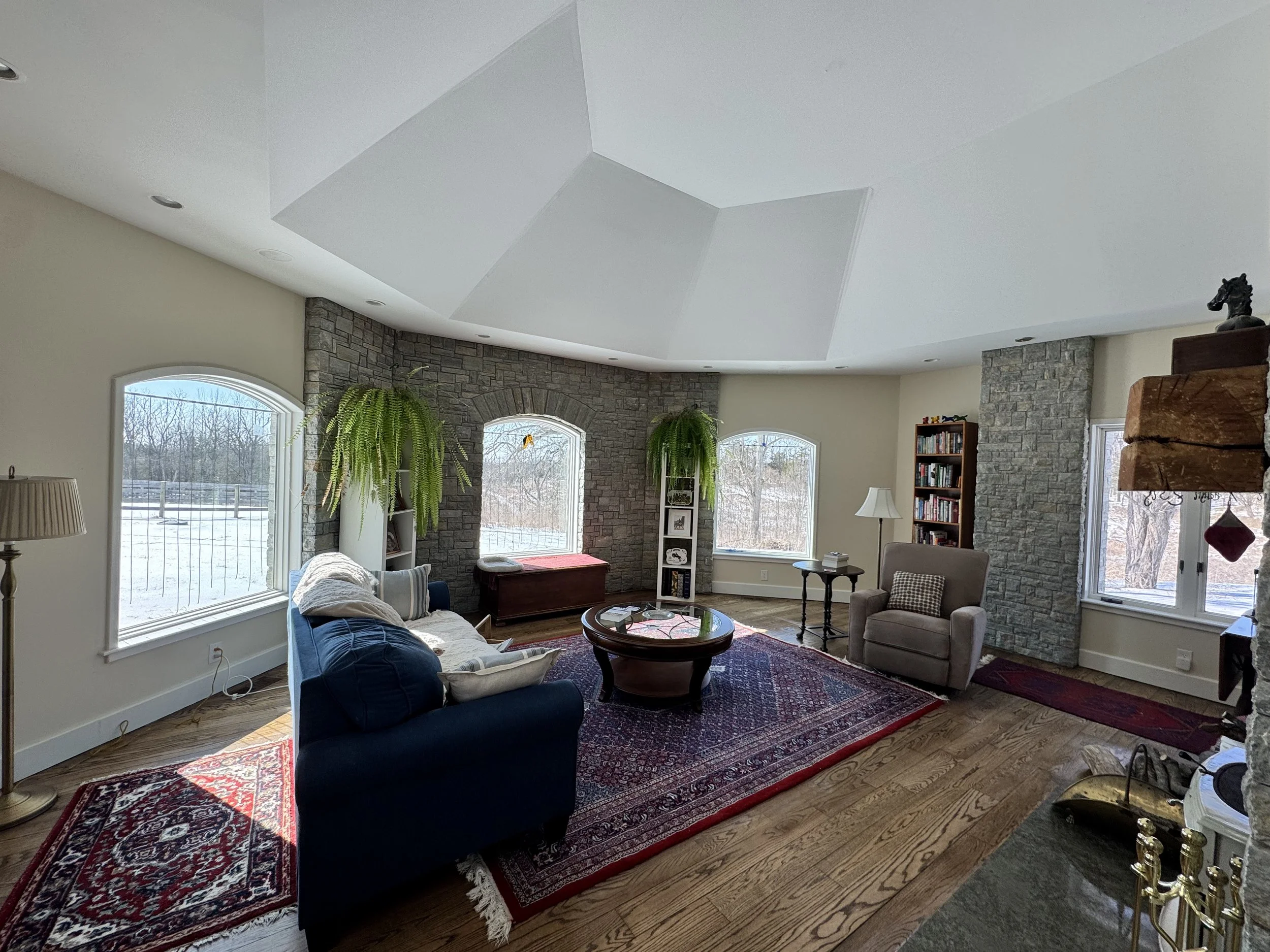 Living room with large windows looking out to a snowy landscape, a blue sofa with pillows, a beige armchair, a round wooden coffee table, a patterned area rug, and some shelves with books and decorative items.