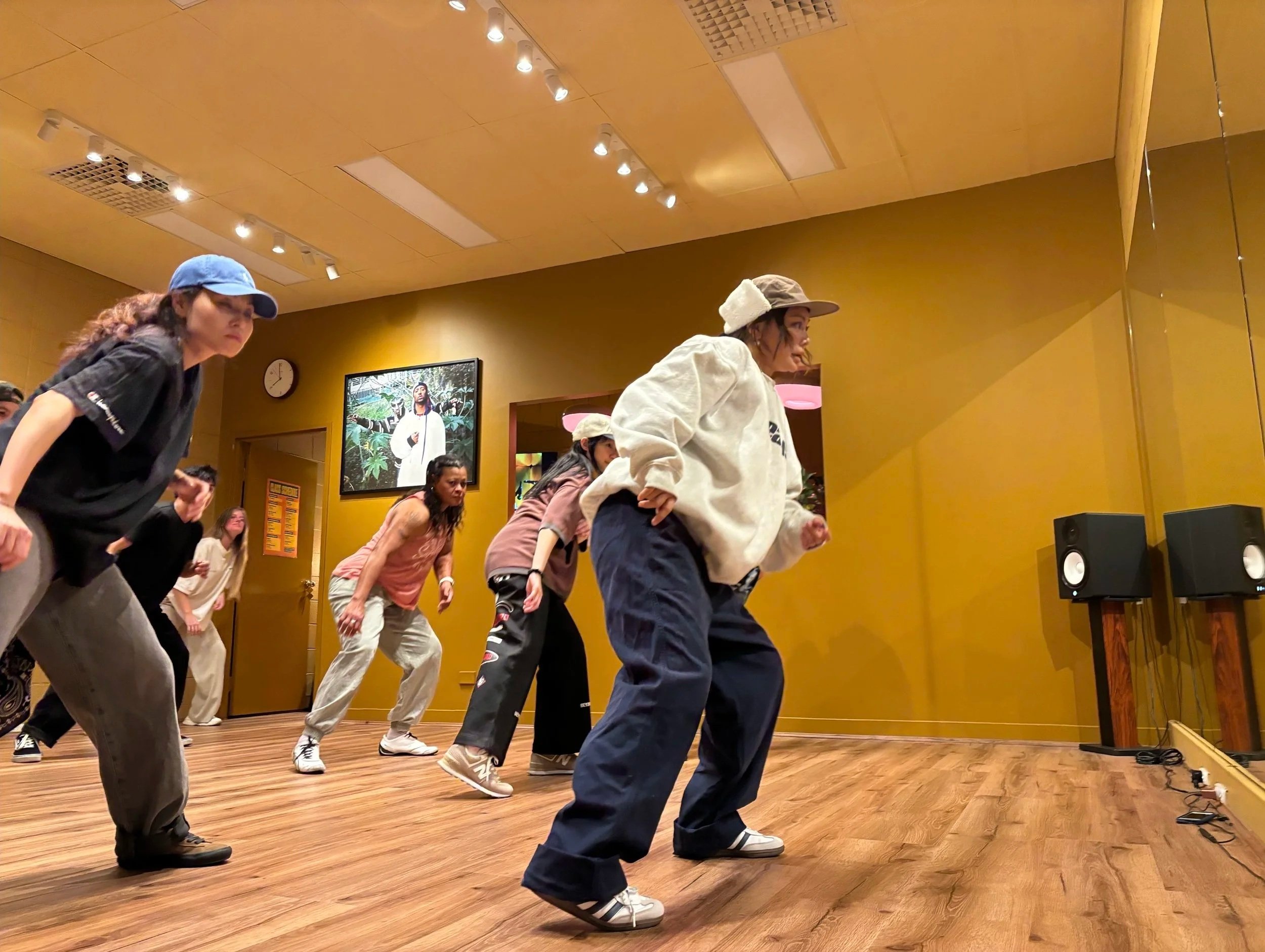Group of men and women dancing in a dance studio with wooden floors and yellow walls. It's a Hip Hop class.
