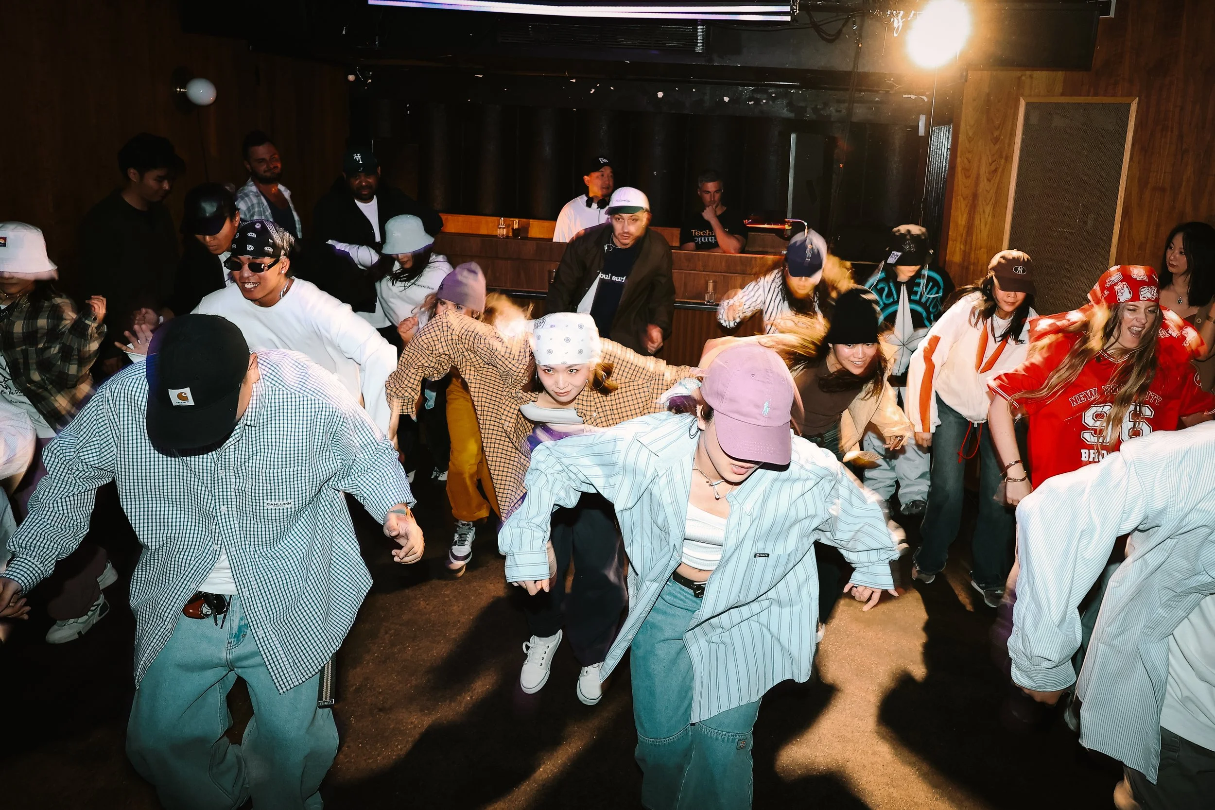 Group of people dancing in a dimly lit indoor venue with wooden walls and a DJ in the background. It's a hip hop party in Perth.