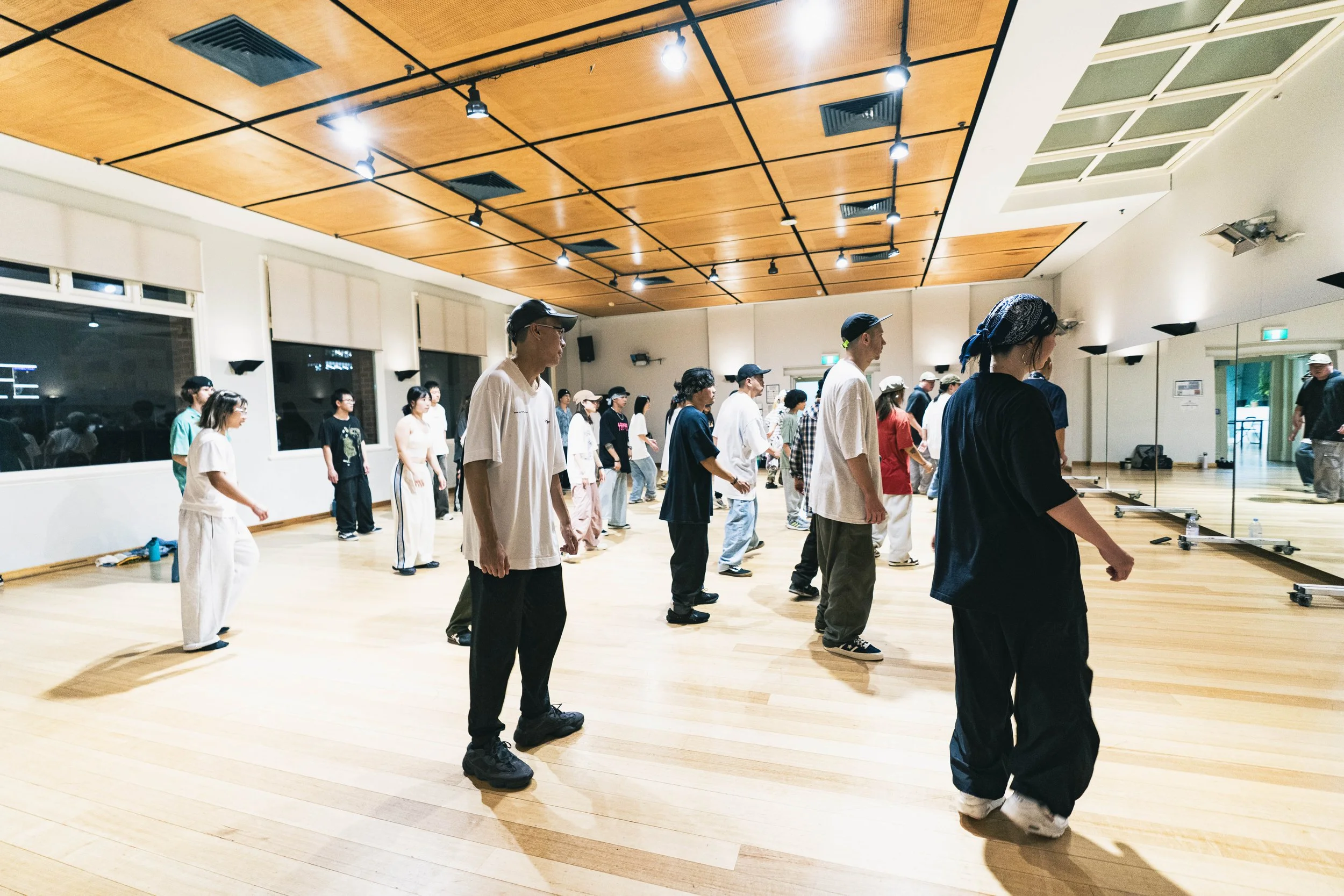 People dancing in a spacious dance studio with wooden floors, large windows, and a mirrored wall.
