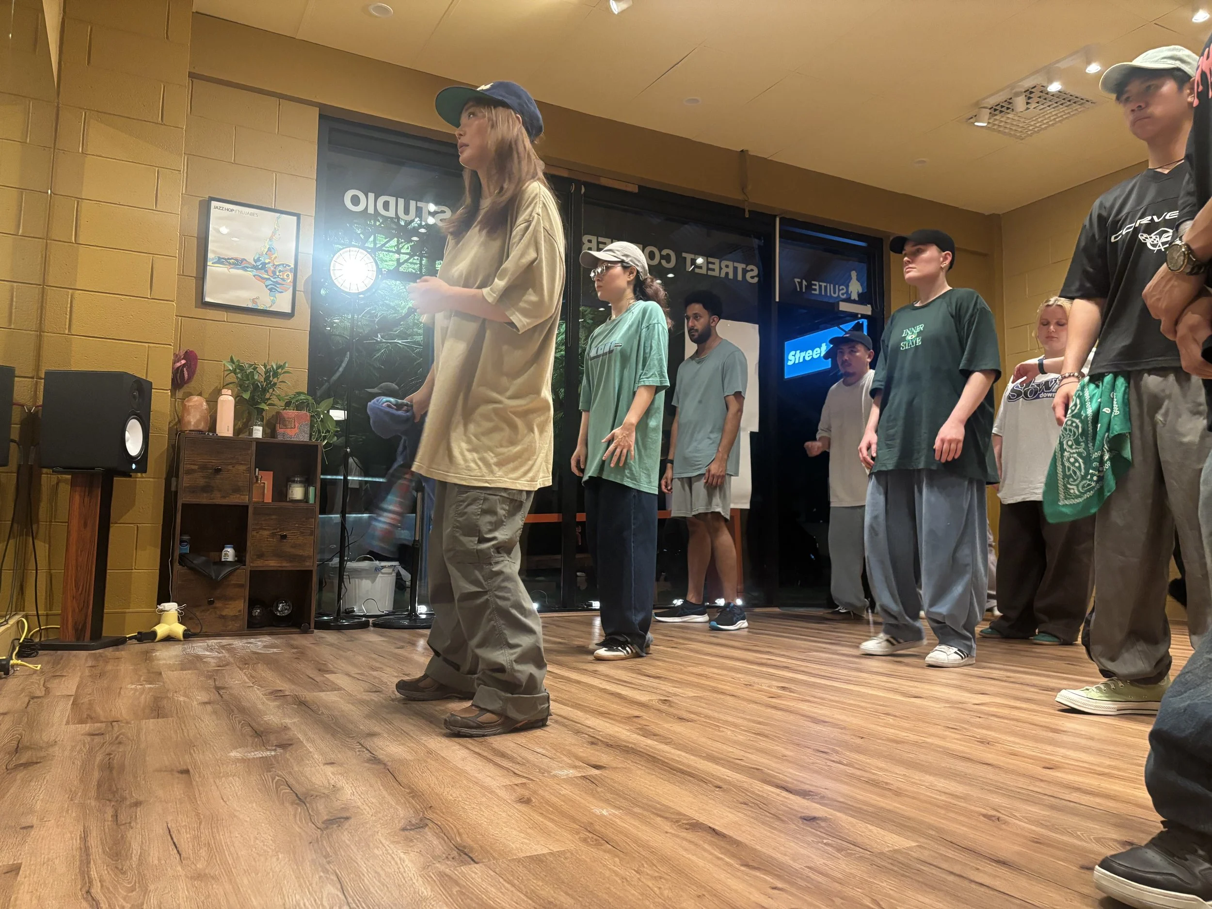 A group of people lined up inside a room with wooden flooring, standing sideways, facing a woman in the front. The woman has long hair, is wearing a beige oversized t-shirt, gray pants, and a blue cap. Some of the people are wearing casual clothes, i