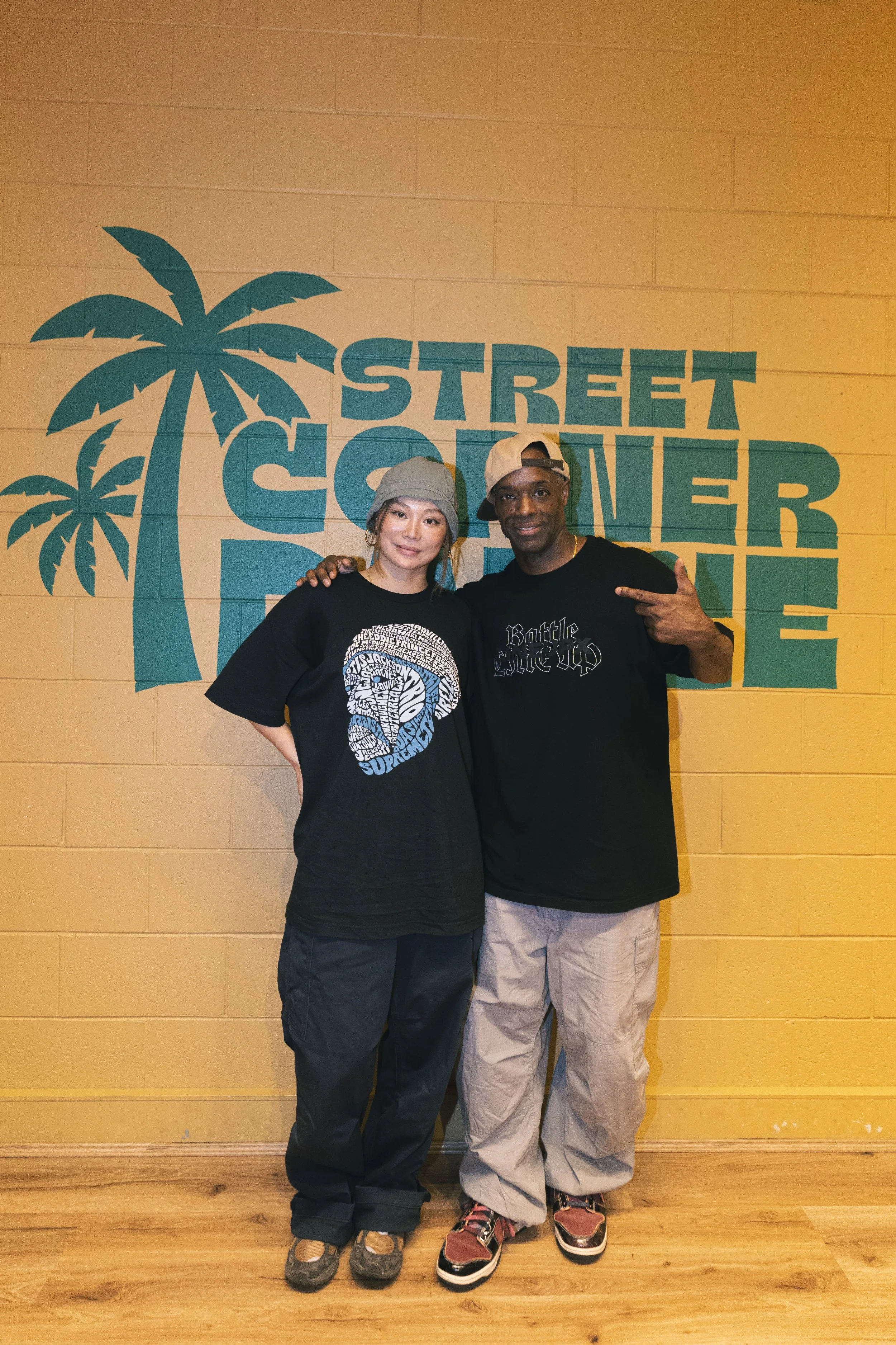 Two individuals standing in front of a yellow wall with turquoise palm trees and the text 'Street Corner Dance' in large letters. They wear Hip Hop Clothes.