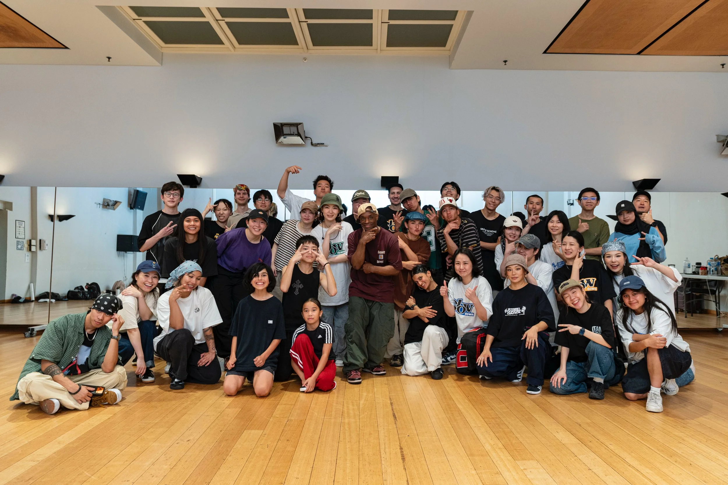 A large group of diverse people posing together in street corner dance studio in Perth with a wooden floor and mirrors, some making gestures and smiling.