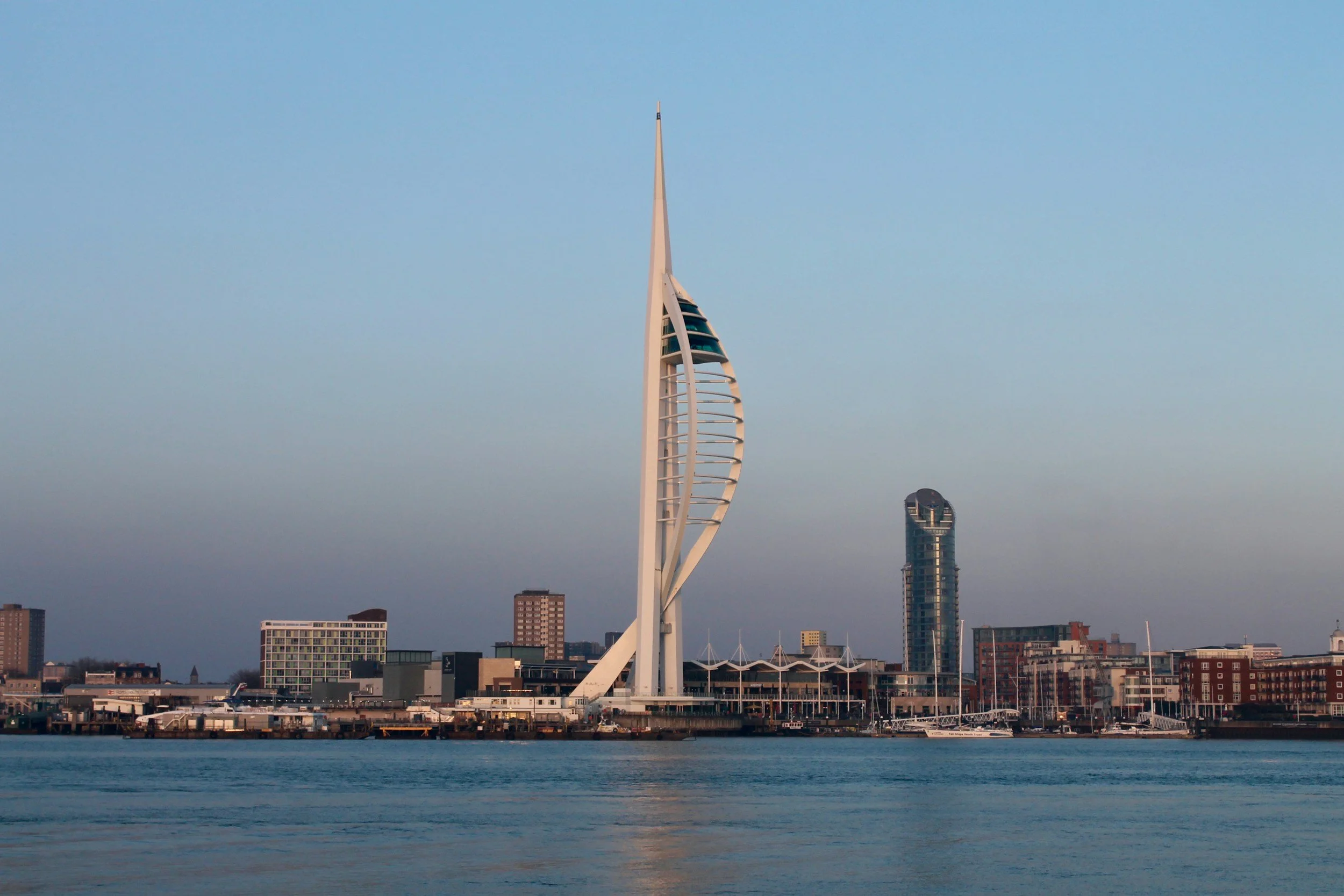 Skyline of a city with a distinctive sail-shaped skyscraper - the Spinnaker Tower, a curved glass high-rise, and other buildings along the waterfront.