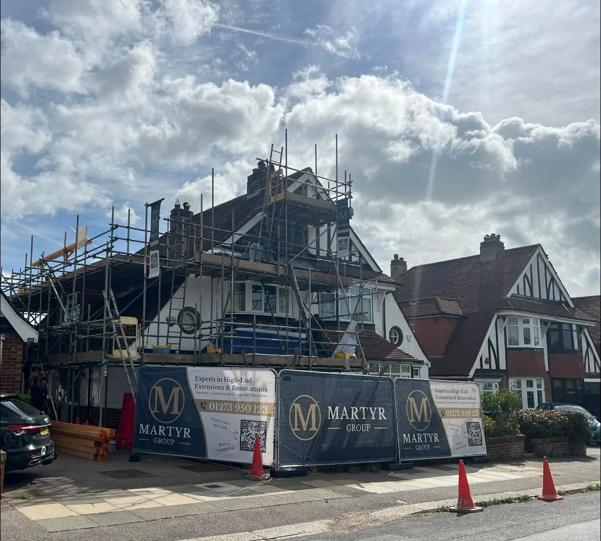 Residential house under renovation with scaffolding around the roof, advertising banners for Martyr Group, and orange traffic cones on the street.