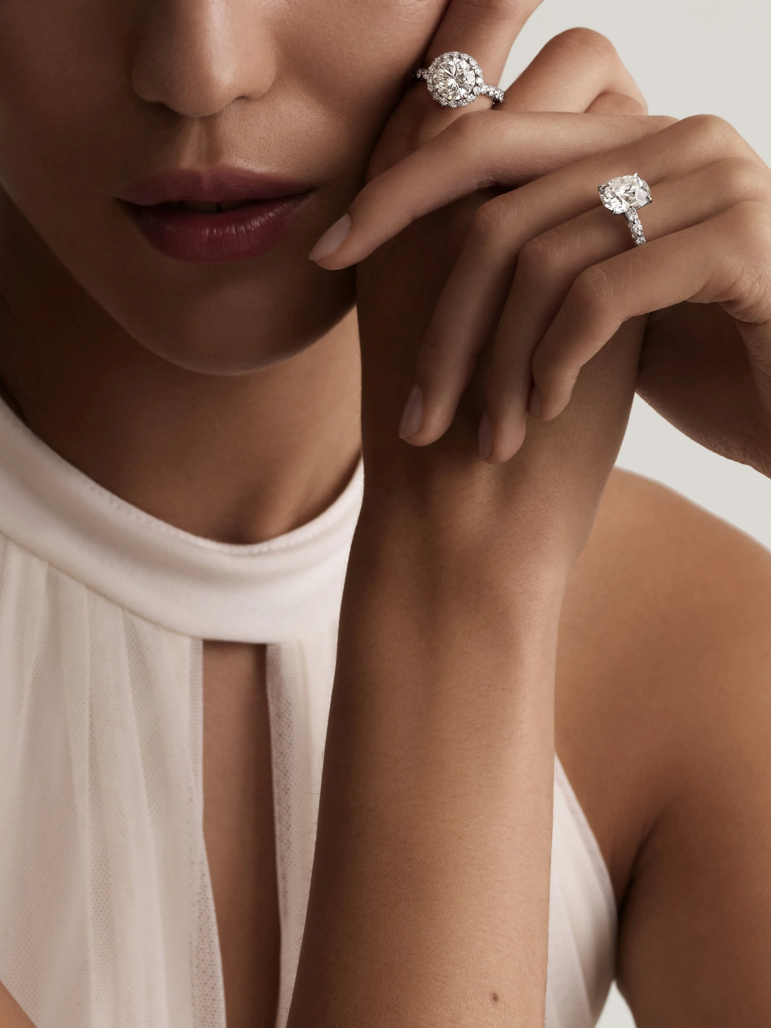 Close-up of a woman wearing diamond rings on her left hand, gently touching her face. The background is plain and light-colored, and the woman is dressed in a white sleeveless top.