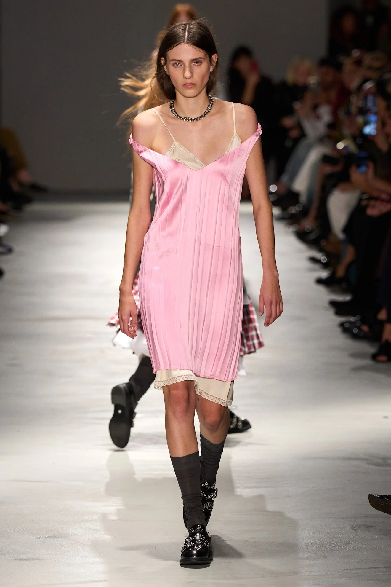 A female model walking down a runway during a fashion show, wearing a pink dress over a beige slip and black shoes with socks.