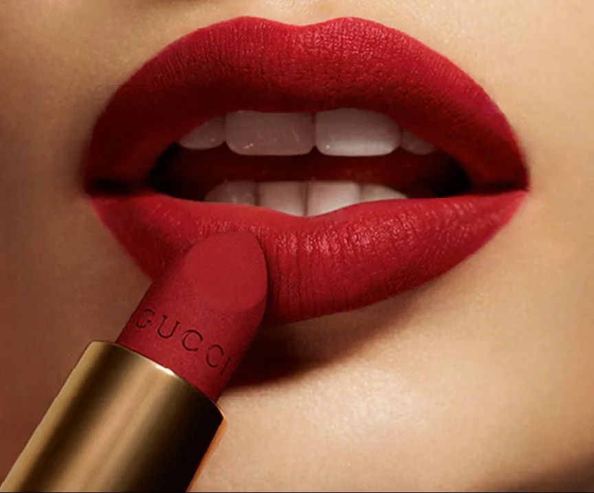 Close-up of a person's lips being painted with red lipstick, with the lipstick tube labeled Gucci.