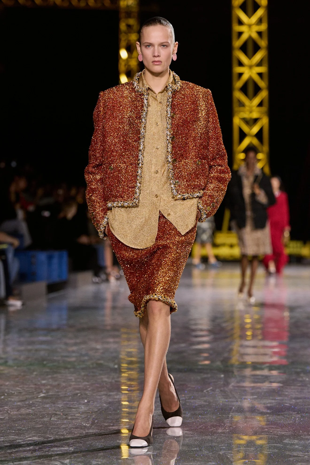 A female runway model walking on Chanel, wearing a glittery red and gold suit with a matching top and pants, accessorized with pink earrings, and black and white high heels, in front of a dark background with yellow stage equipment.