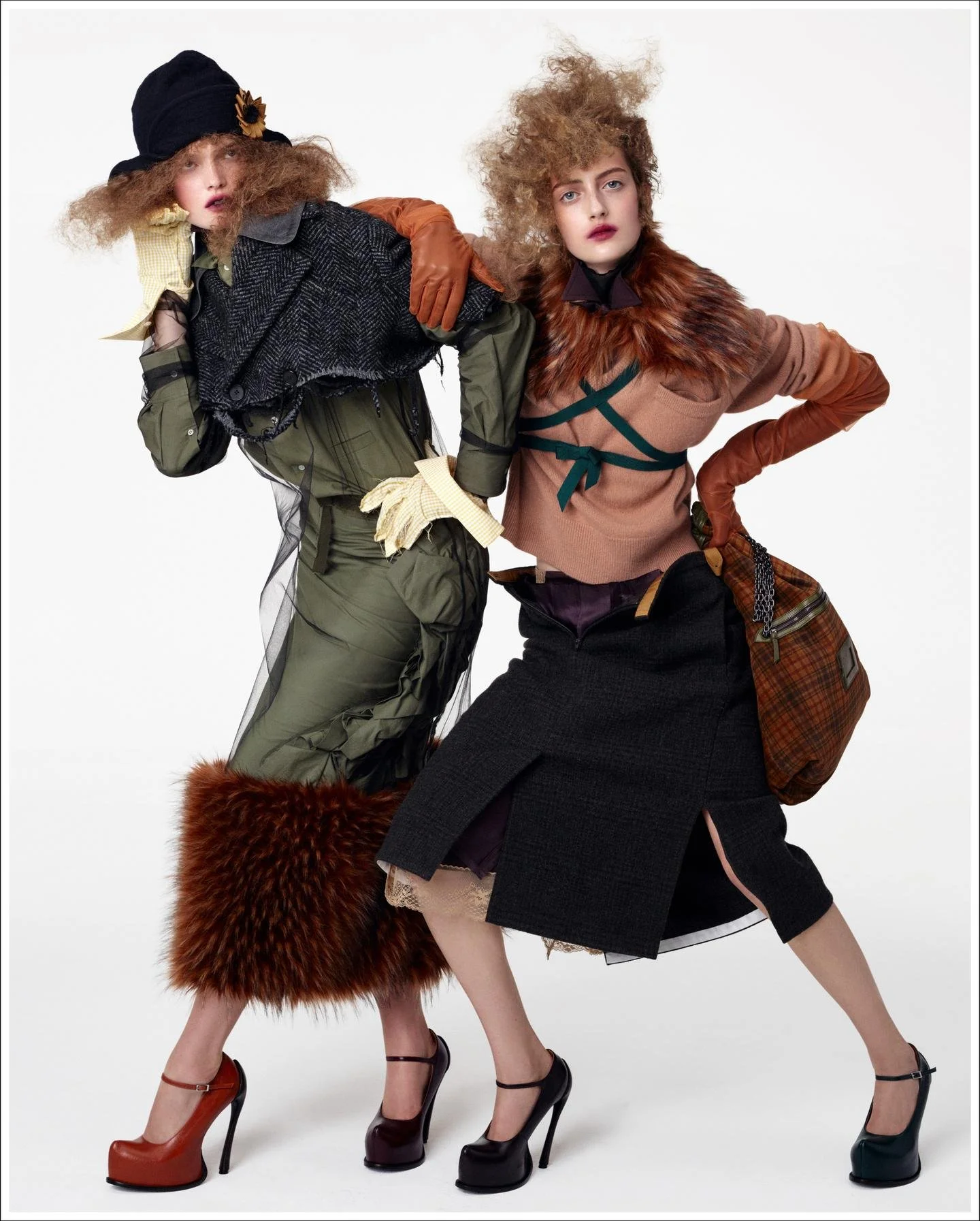Two women with curly, voluminous hair posing in eclectic fashion outfits. One wears a black hat with a flower, a dark blazer, olive-green pants, and high heels. The other wears a fur collar, a beige top, a dark skirt, and high heels. They stand against a plain white background, with one woman resting her hand on her head and the other with her hand on her hip.